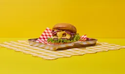 A juicy cheeseburger with lettuce, red onions, and melted cheese served on a metal tray lined with red-and-white checkered paper, set on a yellow gingham cloth and bright yellow background.
