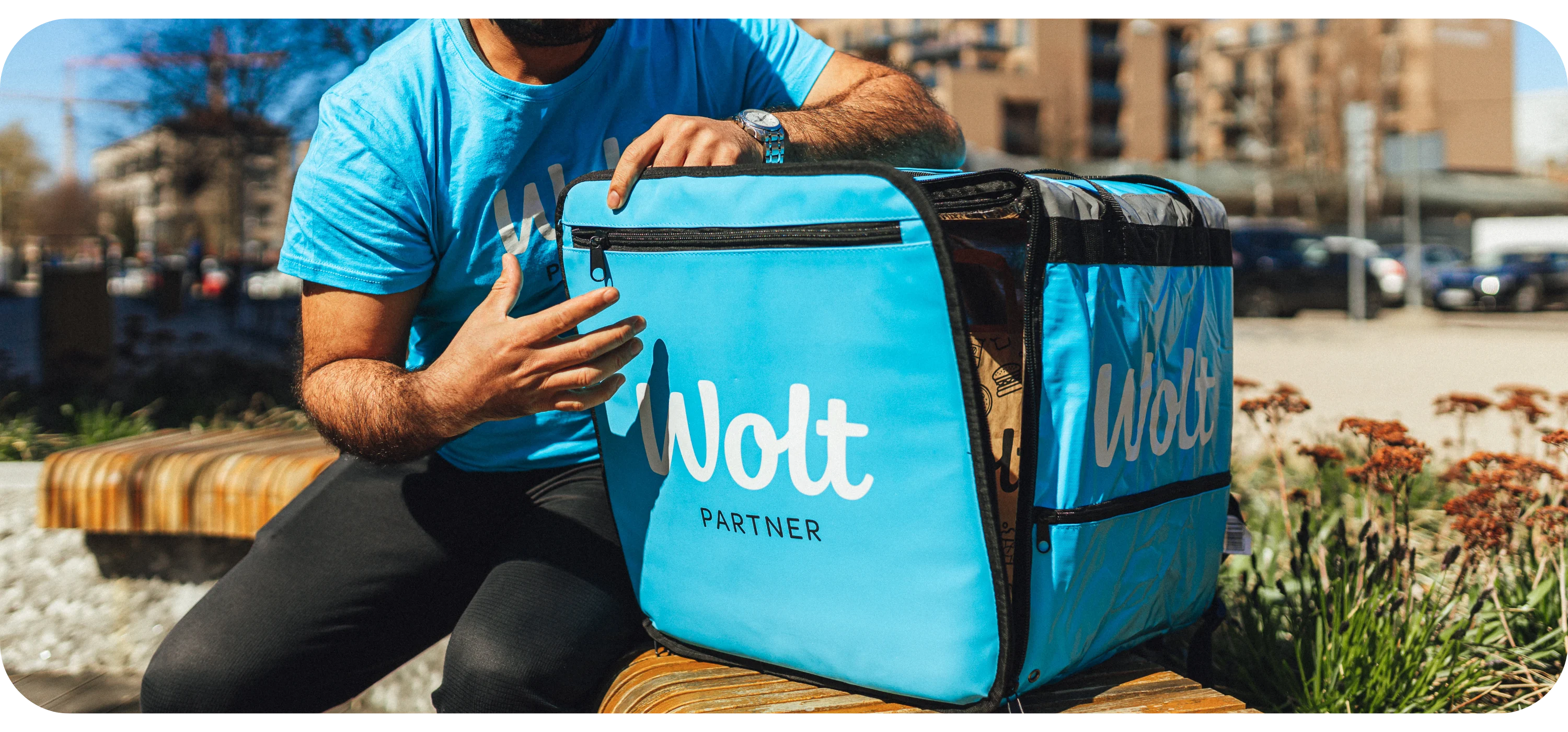 A person in a Wolt shirt sits on a bench holding a blue insulated Wolt Partner delivery bag.