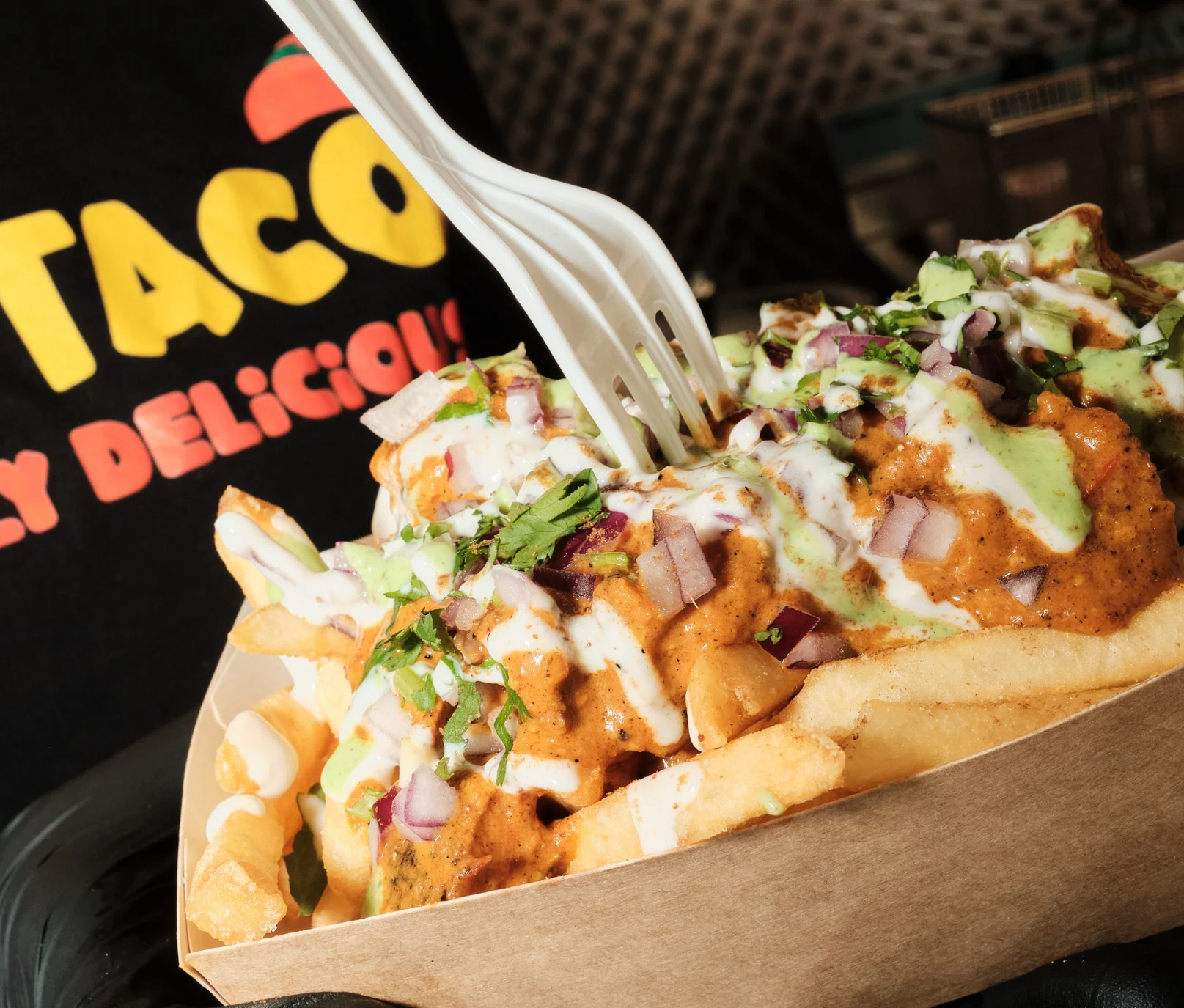 Close-up of Desi Taco’s loaded masala fries topped with creamy sauces, red onions, and cilantro, served in a paper tray.