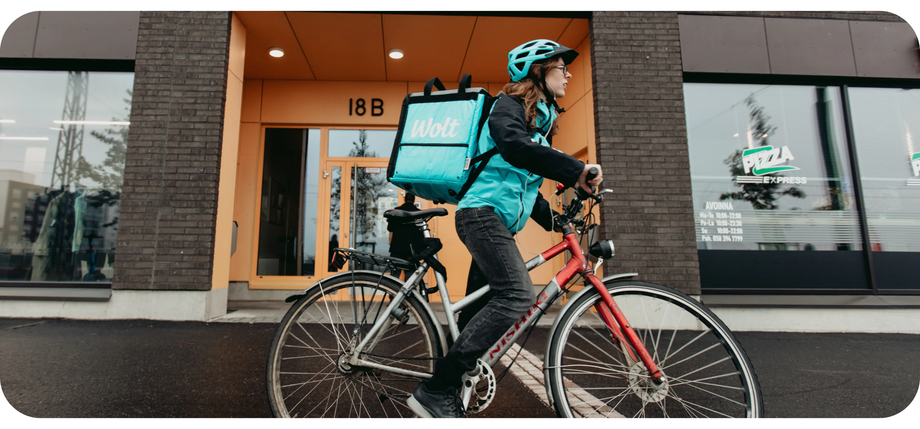 A courier in a helmet rides a bike with a turquoise Wolt delivery backpack past a building entrance and a pizza restaurant.
