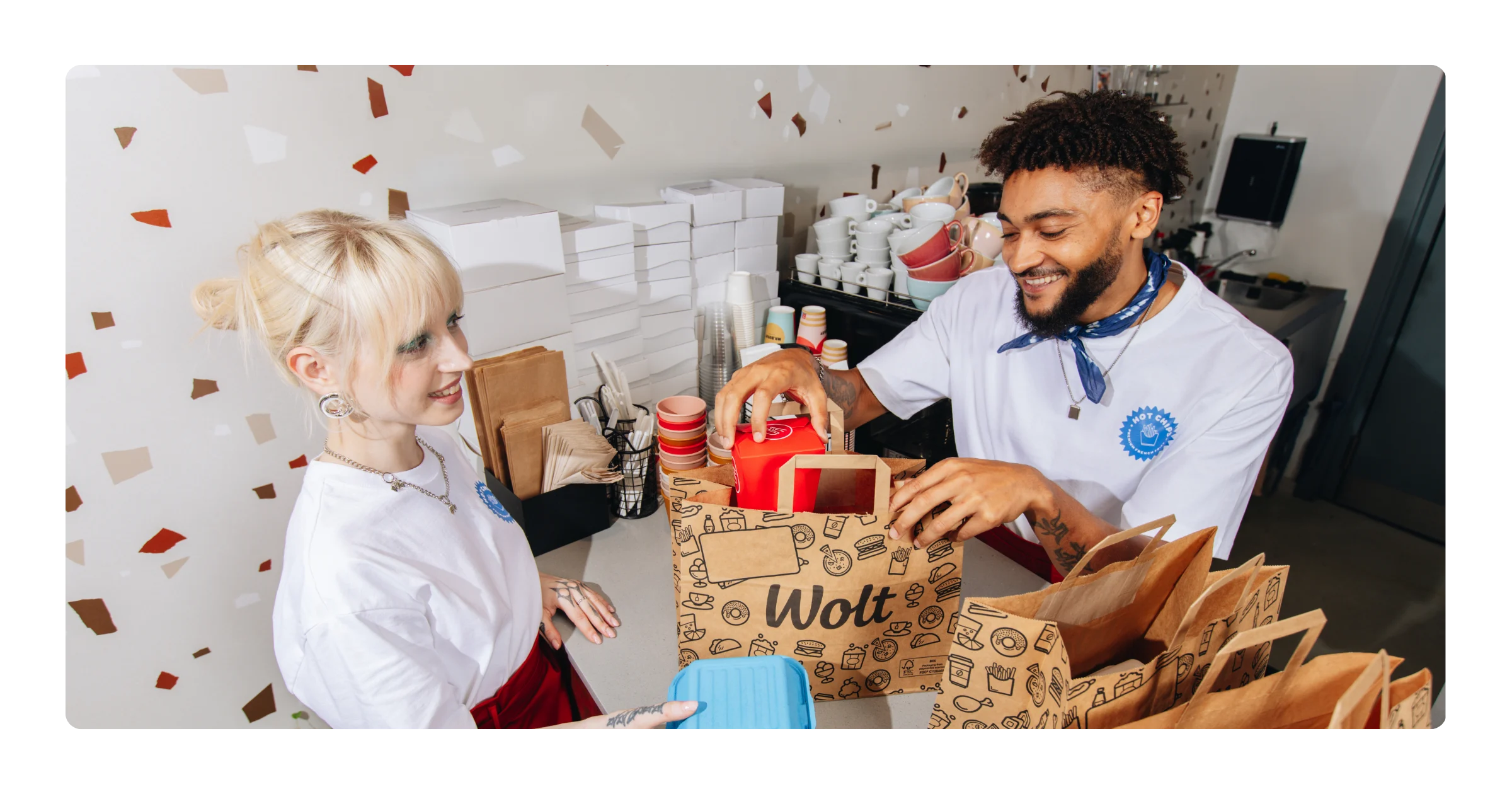 A smiling man and woman pack food containers into paper Wolt bags at a restaurant counter.