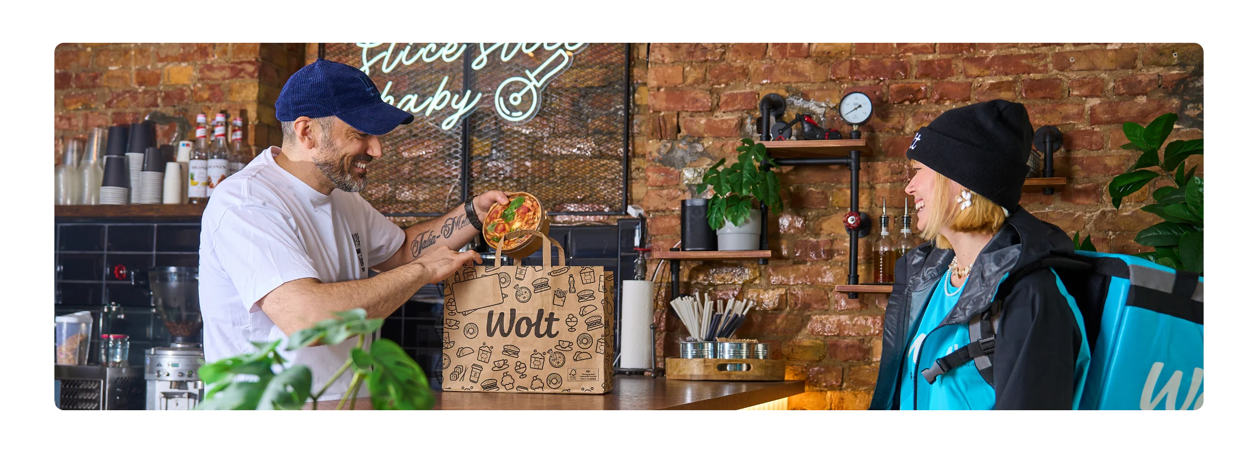 A smiling man in a white t-shirt and navy cap places a pasta bowl into a Wolt paper bag at a restaurant counter, while a Wolt courier in a teal jacket and black beanie smiles back, with a neon sign and brick wall behind them