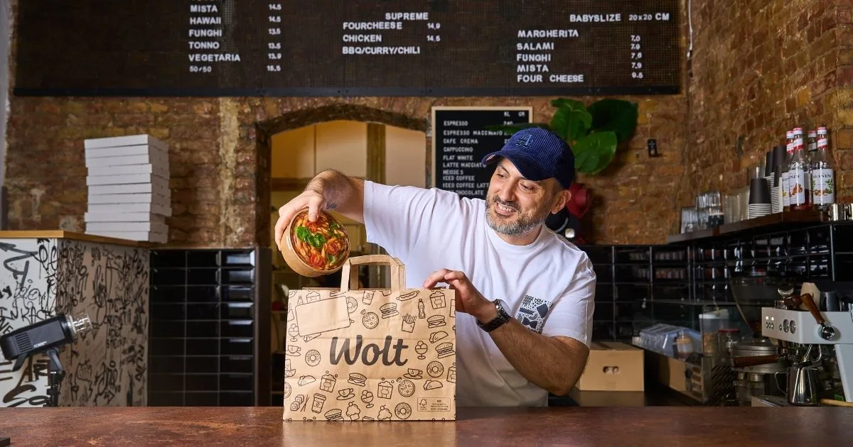 A restaurant owner places a fresh meal into a Wolt delivery bag at the counter, ready for pickup by a courier partner