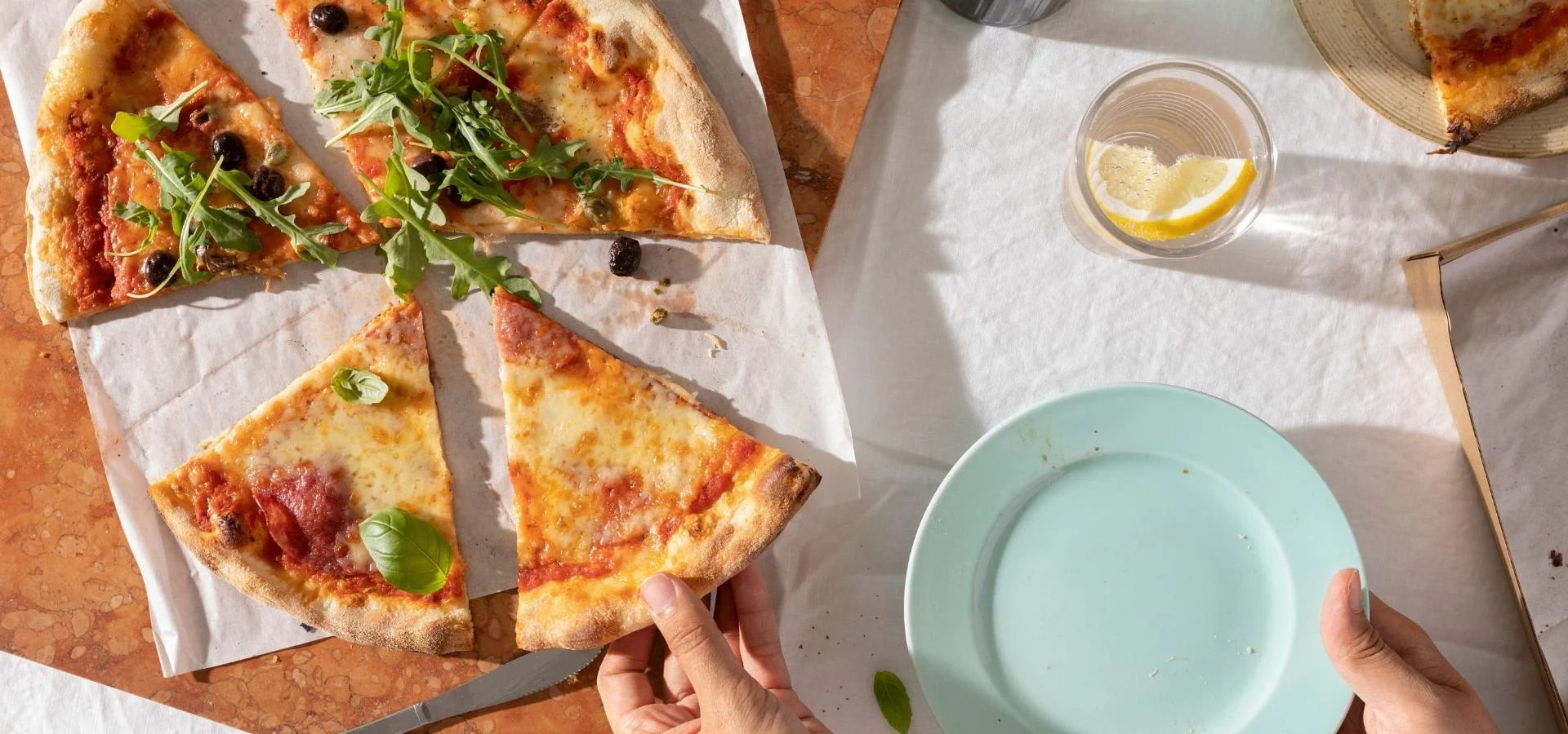  Eine geschnittene Pizza mit Rucola, Oliven und Basilikum auf Backpapier, eine Hand nimmt ein Stück, daneben ein leerer hellblauer Teller und ein Glas Wasser mit Zitrone