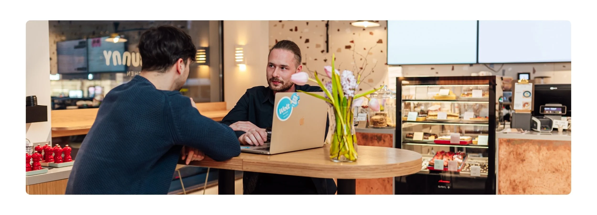 Two men in conversation at a patisserie counter, one working on a laptop with a Wolt sticker, beside a vase of tulips, with a pastry display case behind them