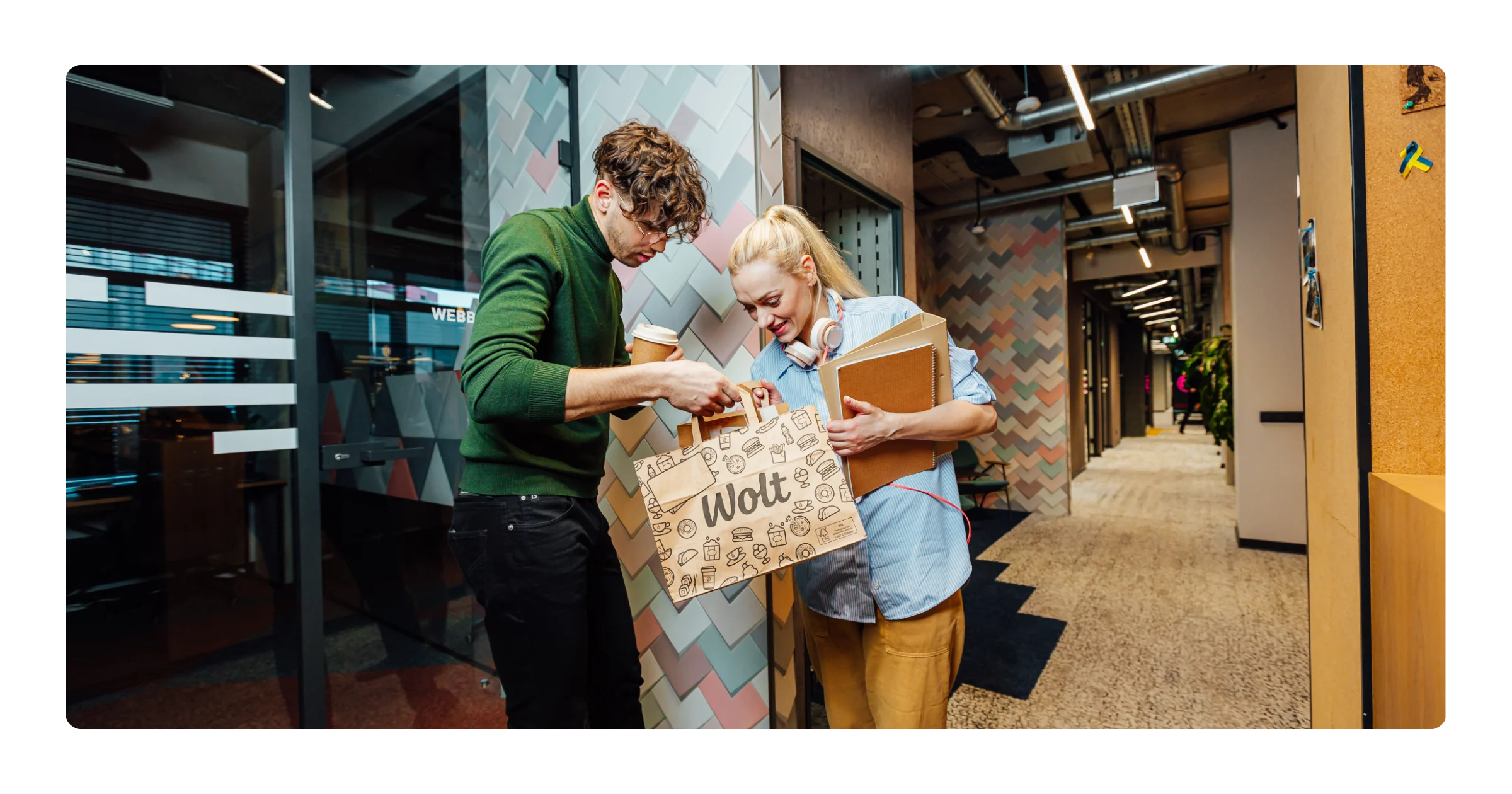 A man and woman look into a paper Wolt bag in an office hallway, the woman holding notebooks and a coffee cup.