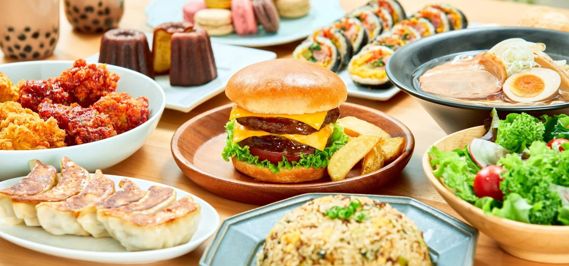 Ein großes Angebot verschiedener Gerichte auf einem Holztisch, darunter ein Cheeseburger mit Pommes, frittiertes Hähnchen, gebratene Teigtaschen, eine Ramen-Schale, Sushi-Rollen, gebratener Reis, ein grüner Salat, Macarons und Bubble Tea