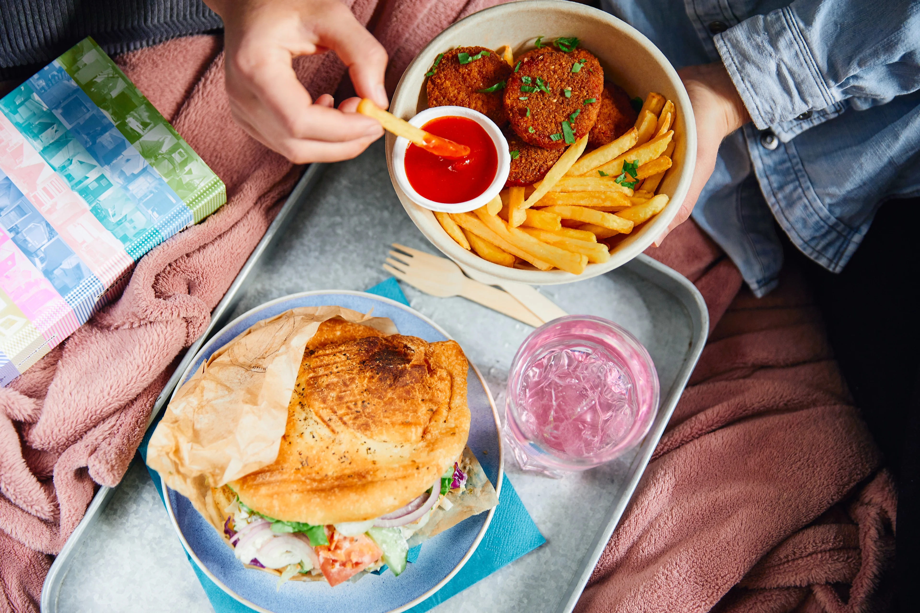 Eine Hand tunkt eine Pommes in Ketchup über einem Tablett mit einer Schale panierter Bratlinge und Pommes, einem Fladenbrot-Sandwich mit Salat auf einem blauen Teller und einem Glas Eiswasser