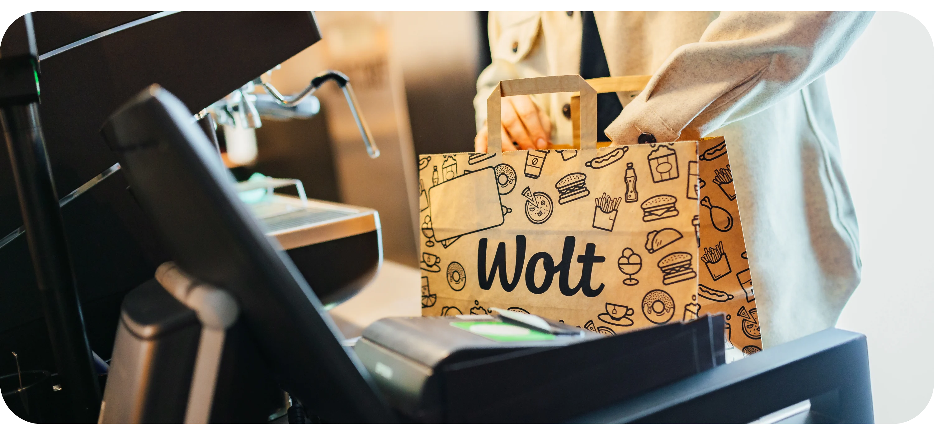 A paper Wolt delivery bag sits on a restaurant counter near a card reader, with a person standing behind it.