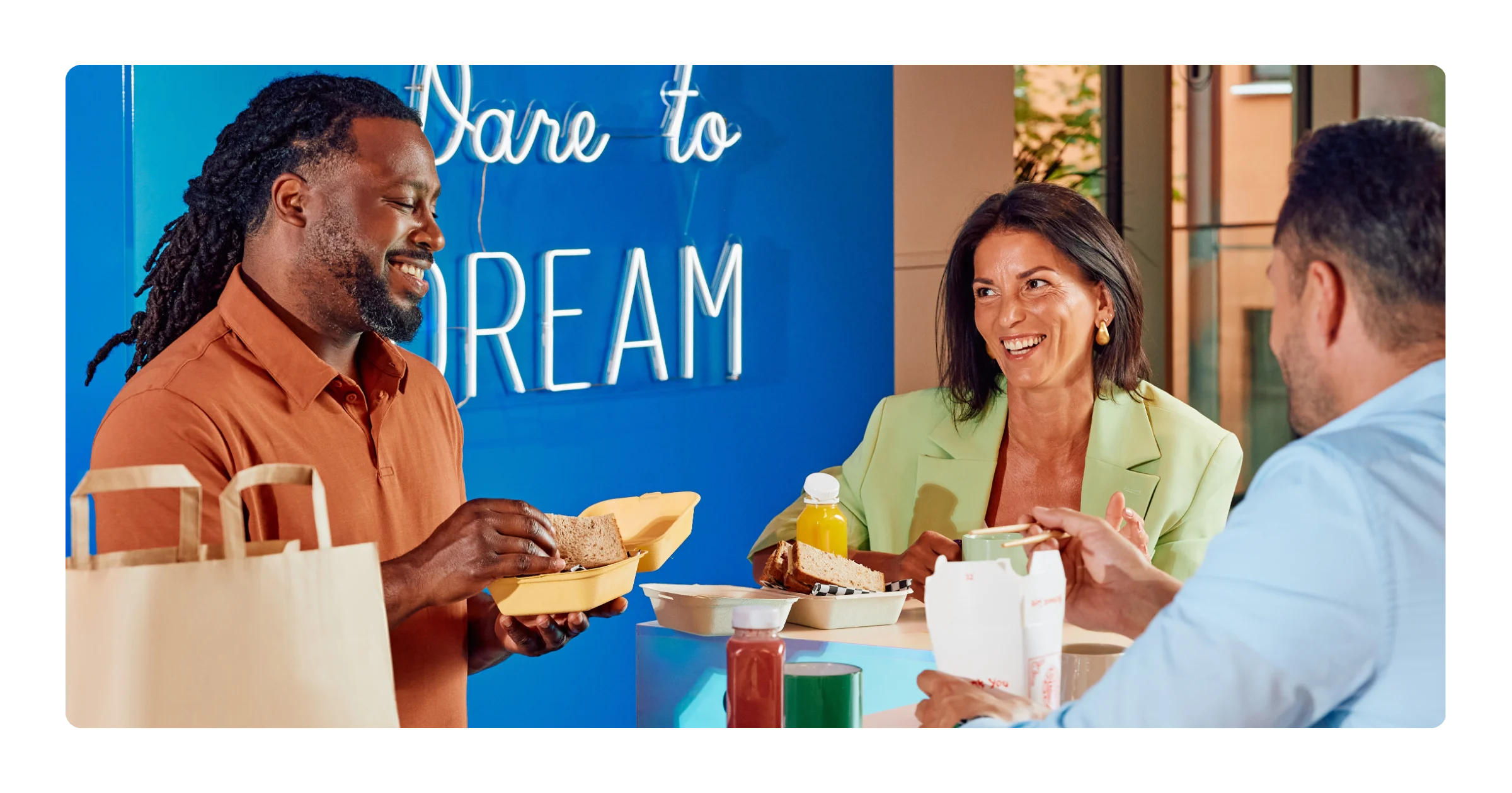 Three people sit at a table sharing takeout food from paper containers, with a paper bag and drinks nearby.
