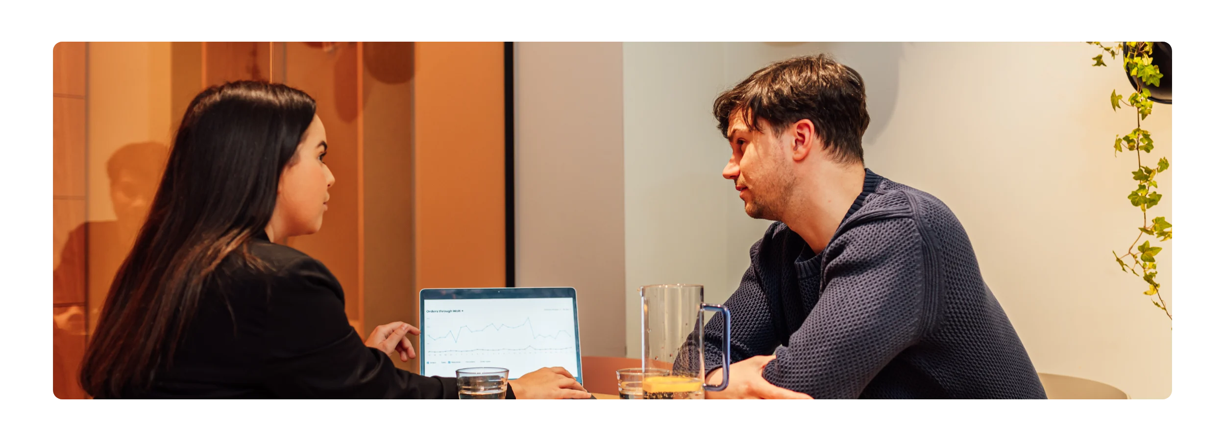 A woman and a man in conversation at a table, with a laptop showing an "Orders through Wolt" line graph, a water pitcher, and glasses between them