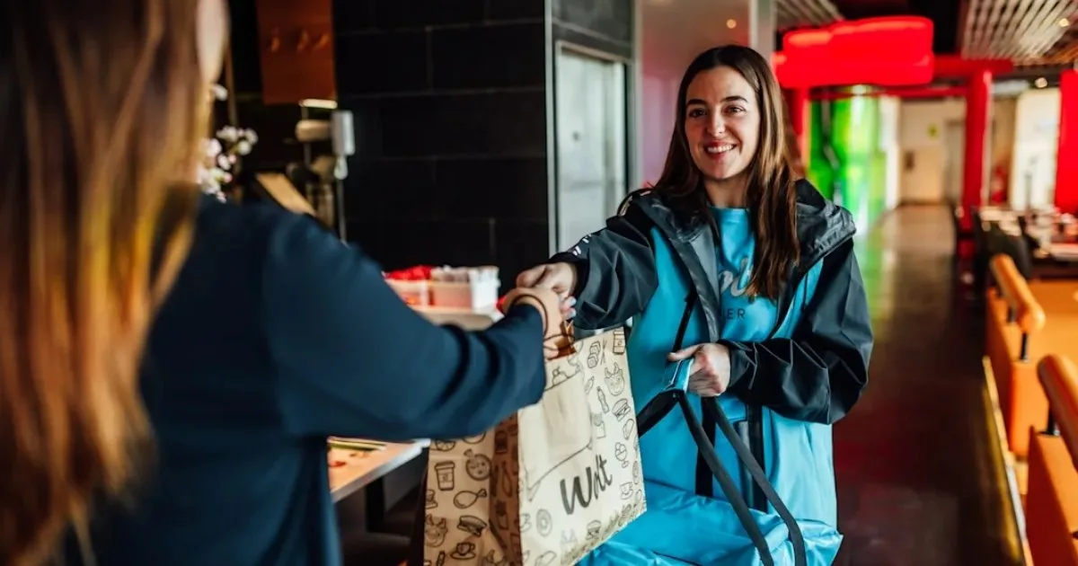 A Wolt courier partner picks up a delivery bag from a restaurant staff member, illustrating how a food delivery platform handles last-mile logistics for restaurants