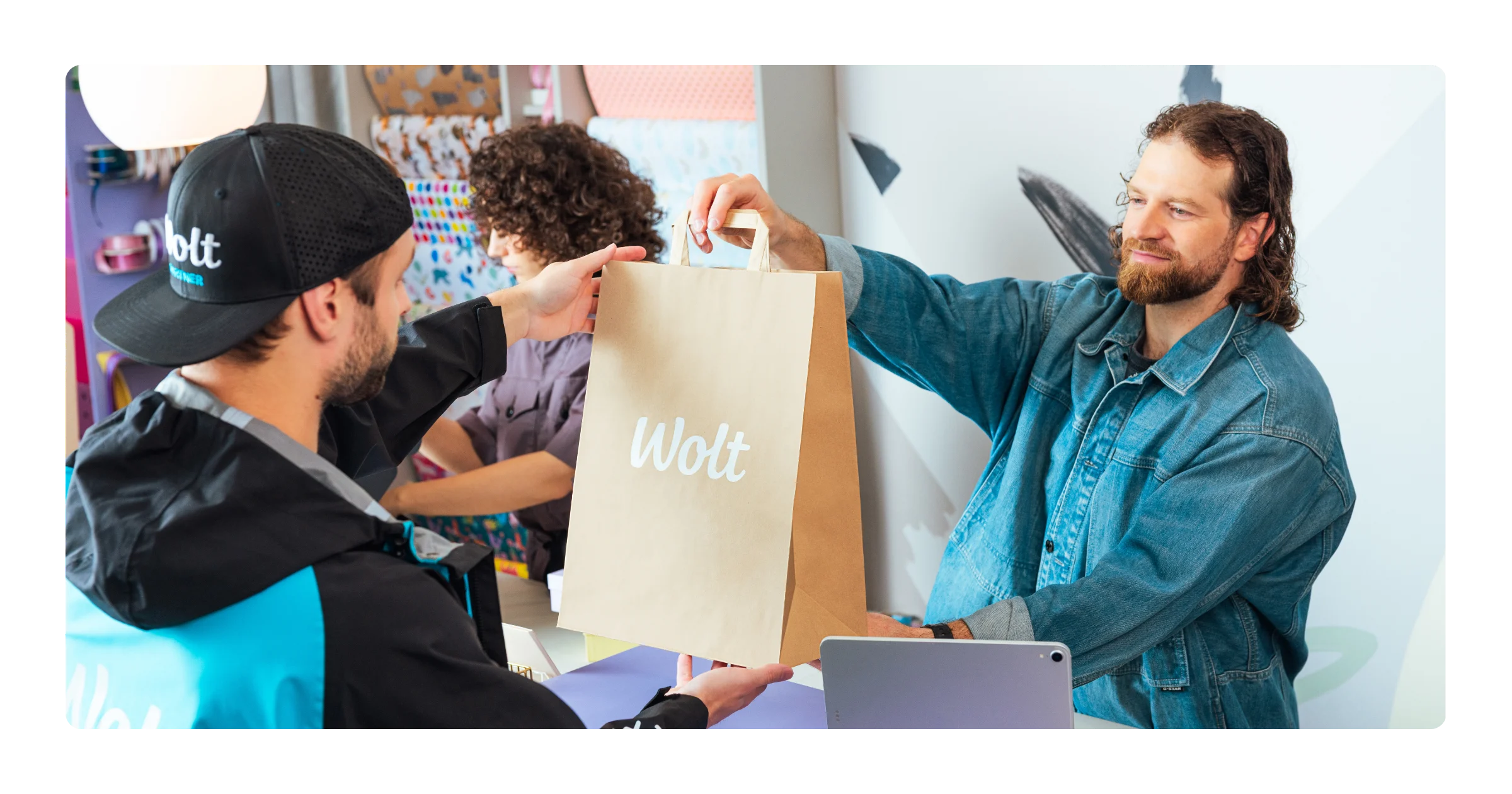 A courier wearing a Wolt cap and jacket hands a paper Wolt bag to a man, with another person nearby.