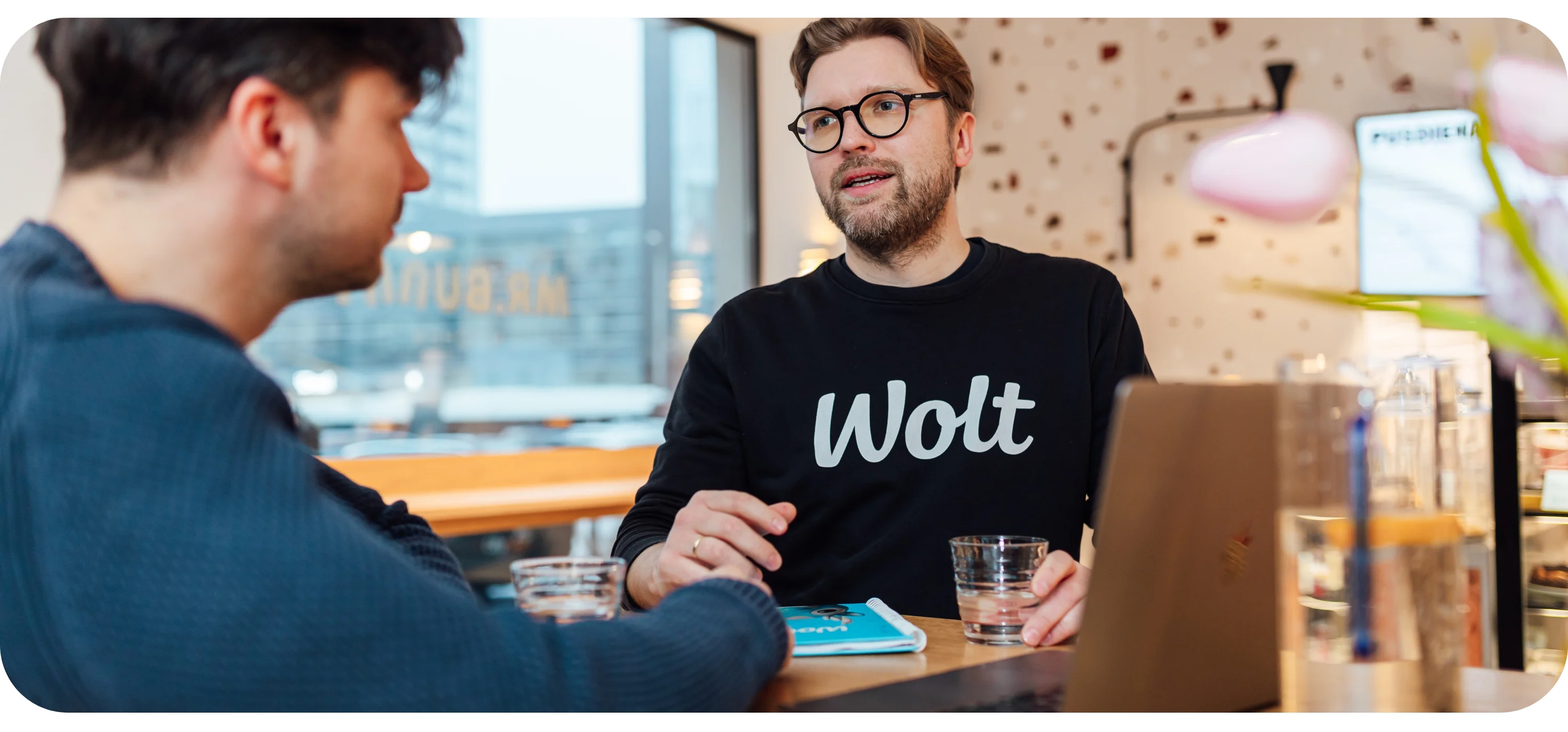 A man in a black Wolt sweatshirt talks with another person across a table