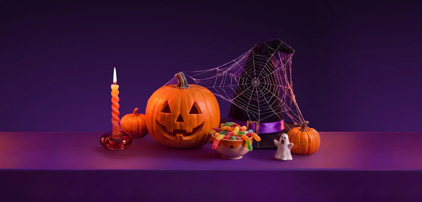 A carved jack-o'-lantern pumpkin and a smaller pumpkin beside a lit spiral candle, a bowl of colorful gummy worm candies, a witch hat draped in spiderweb, and a small ghost figurine, arranged on a purple surface, representing Halloween treats delivered with Wolt.