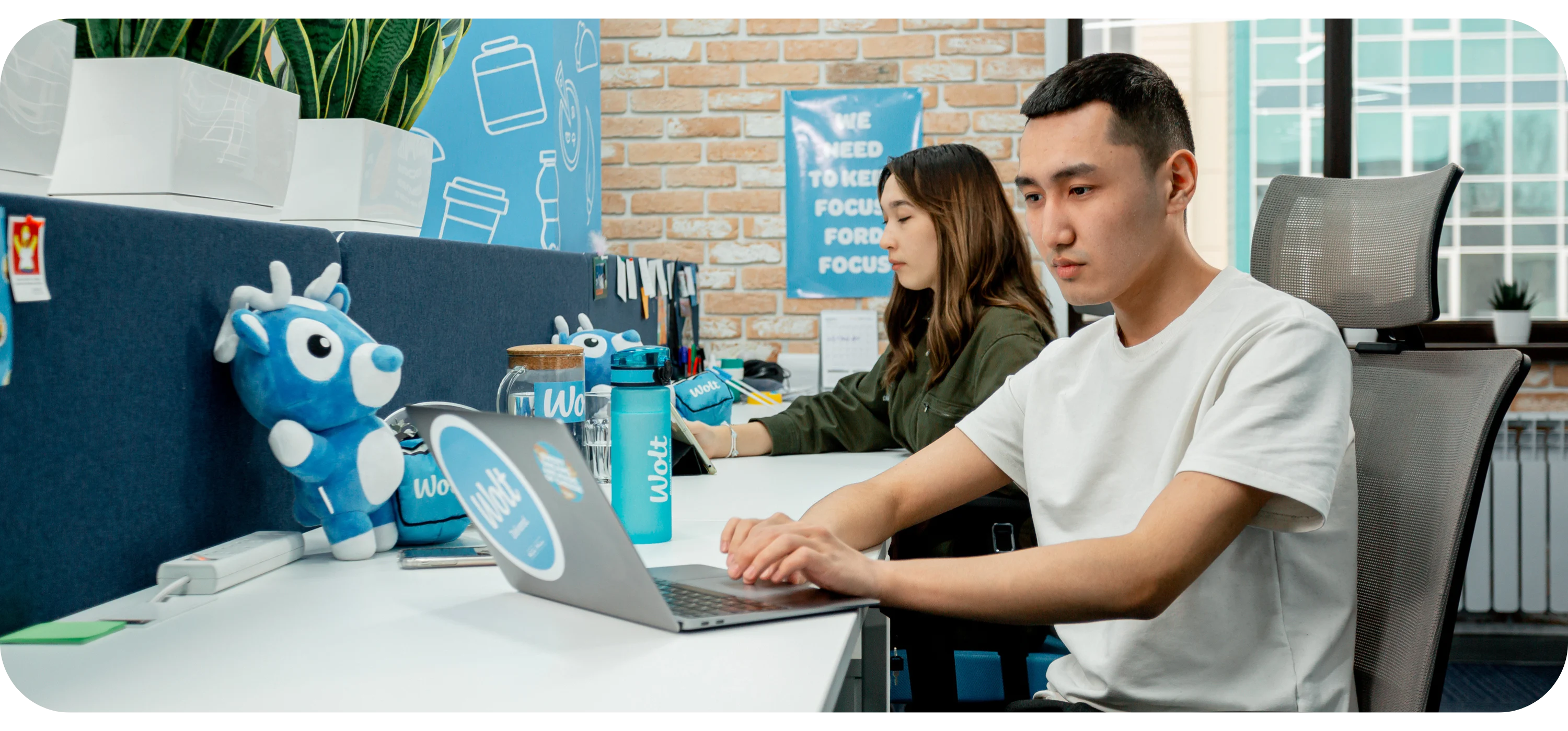 A man types on a laptop with Wolt stickers, beside a woman working, surrounded by Wolt-branded office items.