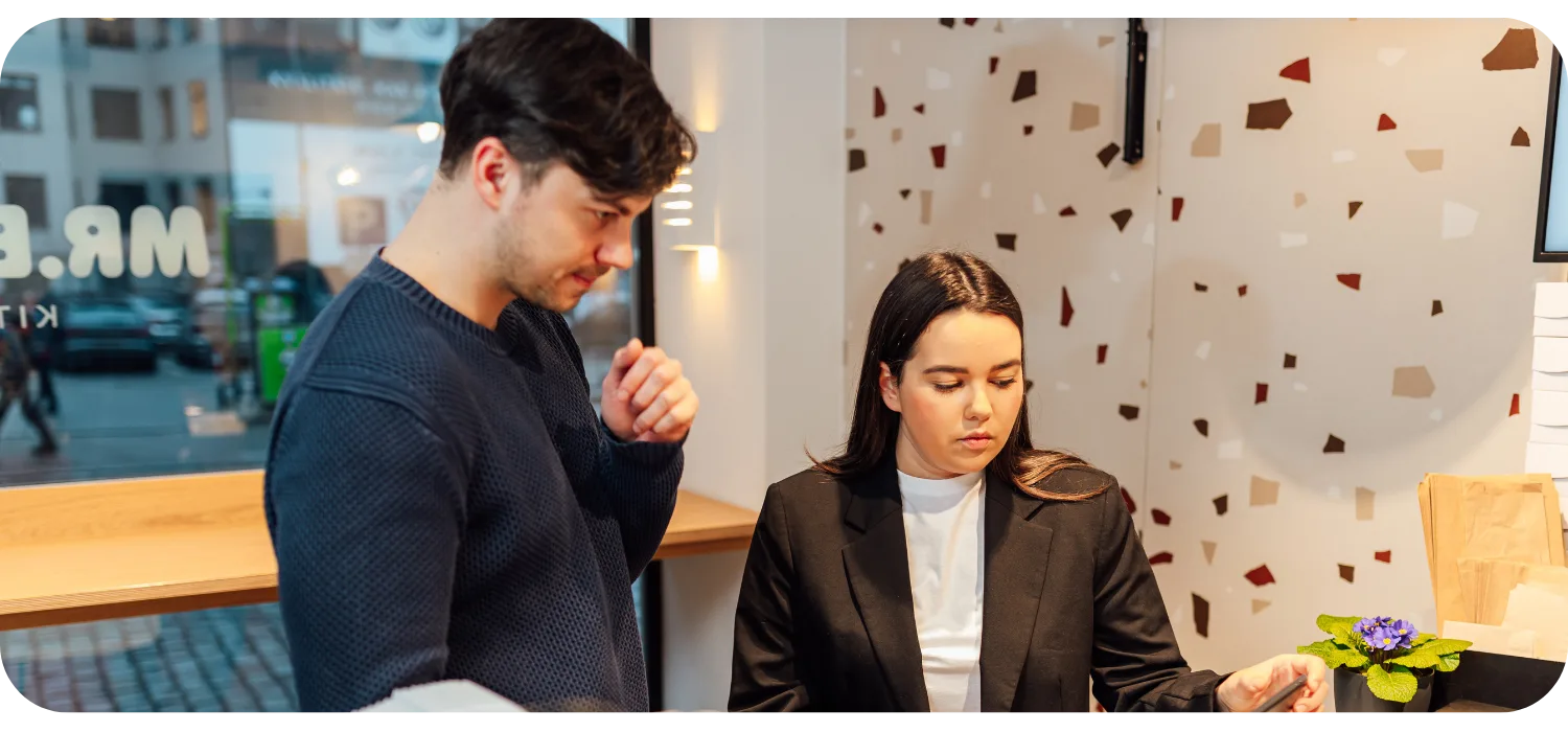 Two restaurant staff members focus on a device at the counter, managing orders during a busy period with takeaway bags ready