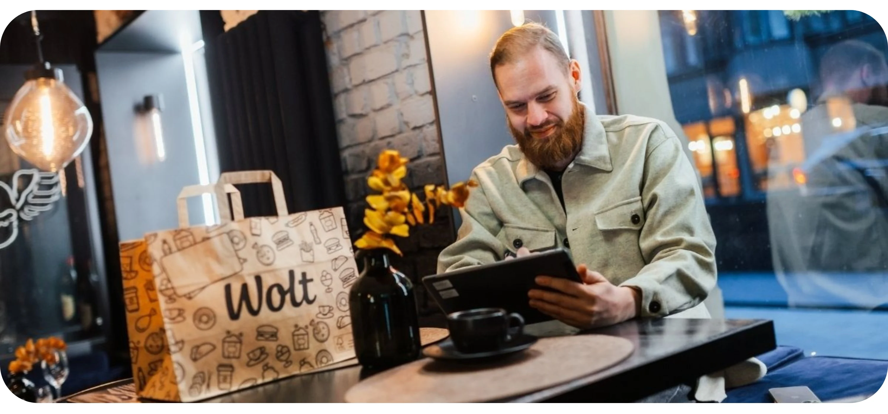 A bearded man reviews figures on a tablet at a café table next to a Wolt paper bag and a coffee
