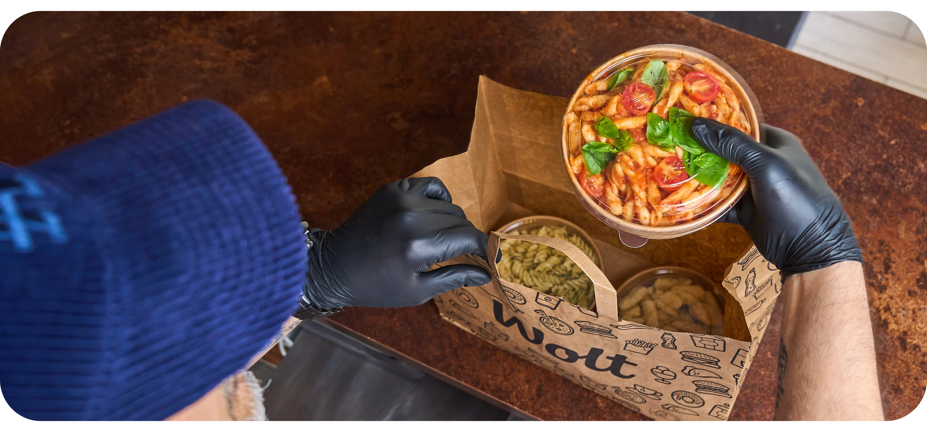 Gloved hands pack a bowl of tomato pasta and two containers of pasta into an open Wolt delivery box.