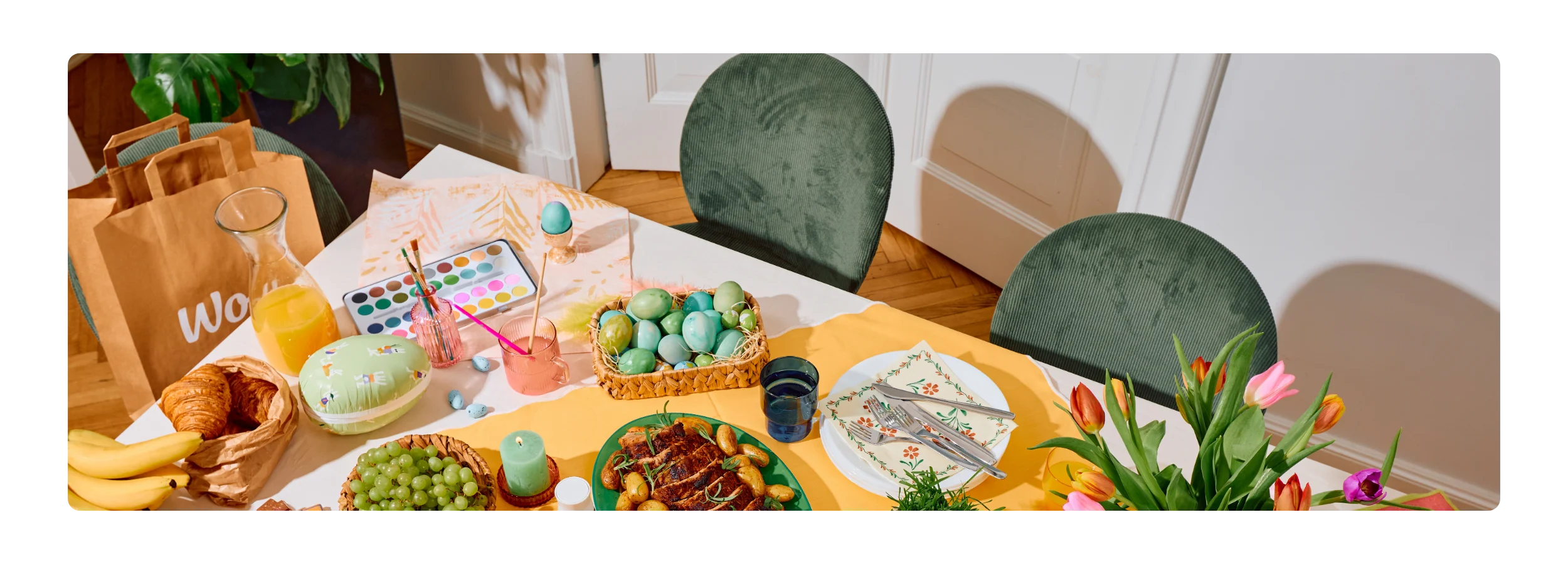 Ein Ostertisch von oben mit einer Wolt-Papiertüte, gefärbten Eiern, einem Wasserfarbkasten, Braten mit Kartoffeln, Croissants, Obst, Saft und Tulpen