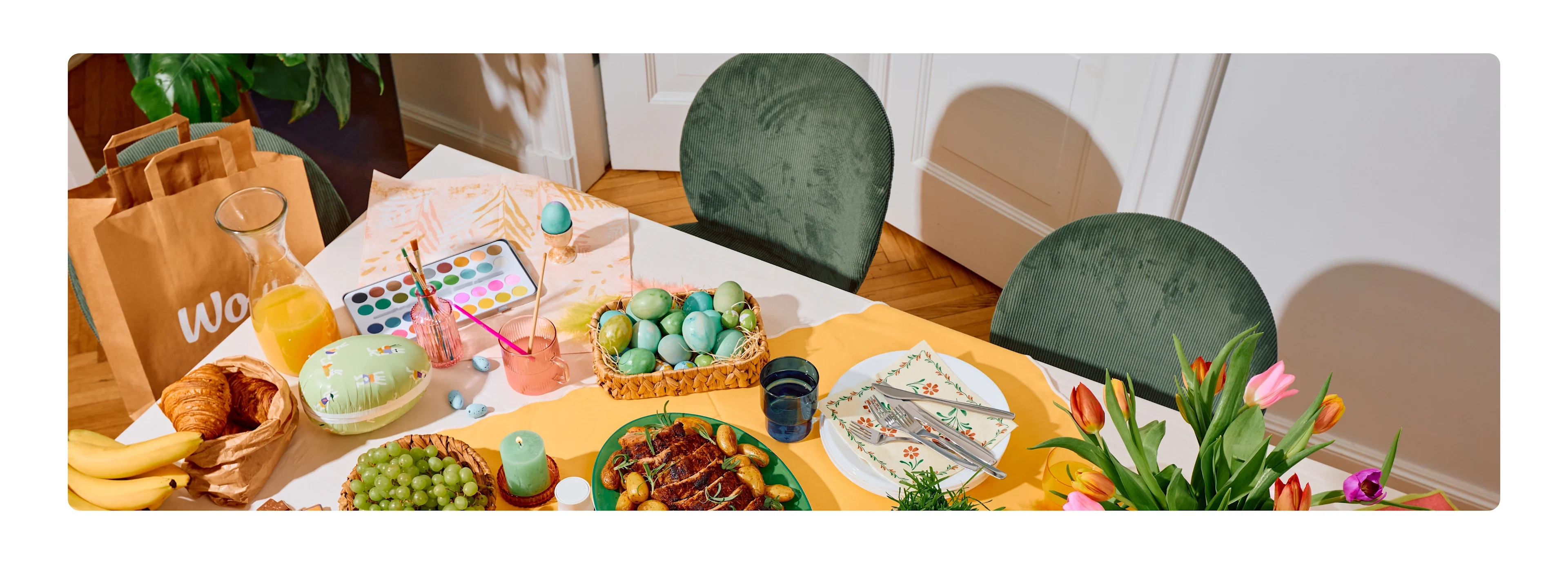 Ein Ostertisch von oben mit einer Wolt-Papiertüte, gefärbten Eiern, einem Wasserfarbkasten, Braten mit Kartoffeln, Croissants, Obst, Saft und Tulpen