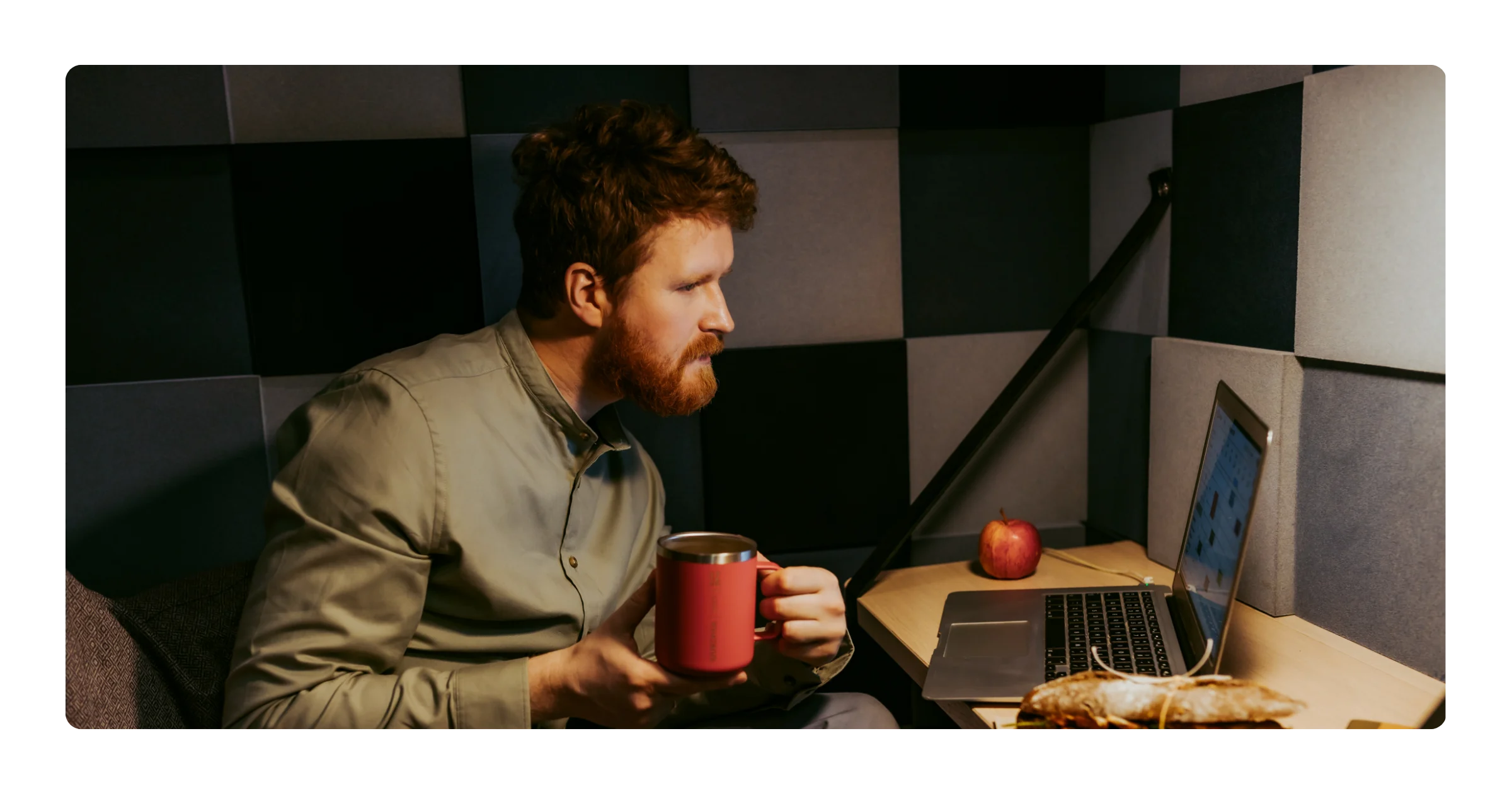 A bearded man sits in a booth holding a red mug, with a laptop, an apple, and a sandwich on the table.