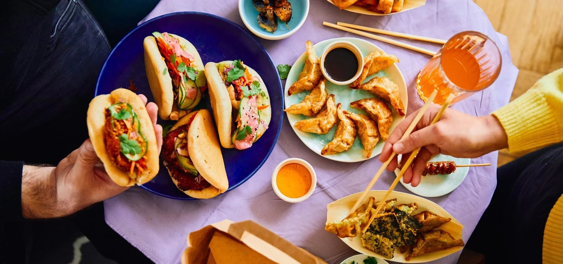 Aufsicht auf zwei Personen, die sich eine Mahlzeit teilen, mit Bao-Buns, frittierten Teigtaschen und Dips, einem Spieß und einem Glas Orangengetränk auf einem Tisch