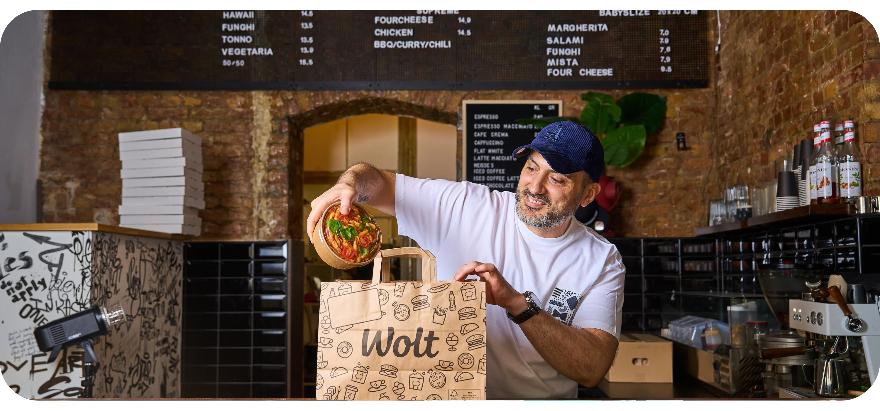 A man behind a counter places a bowl of pasta into a paper Wolt delivery bag, with a pizza menu board on the wall behind him.