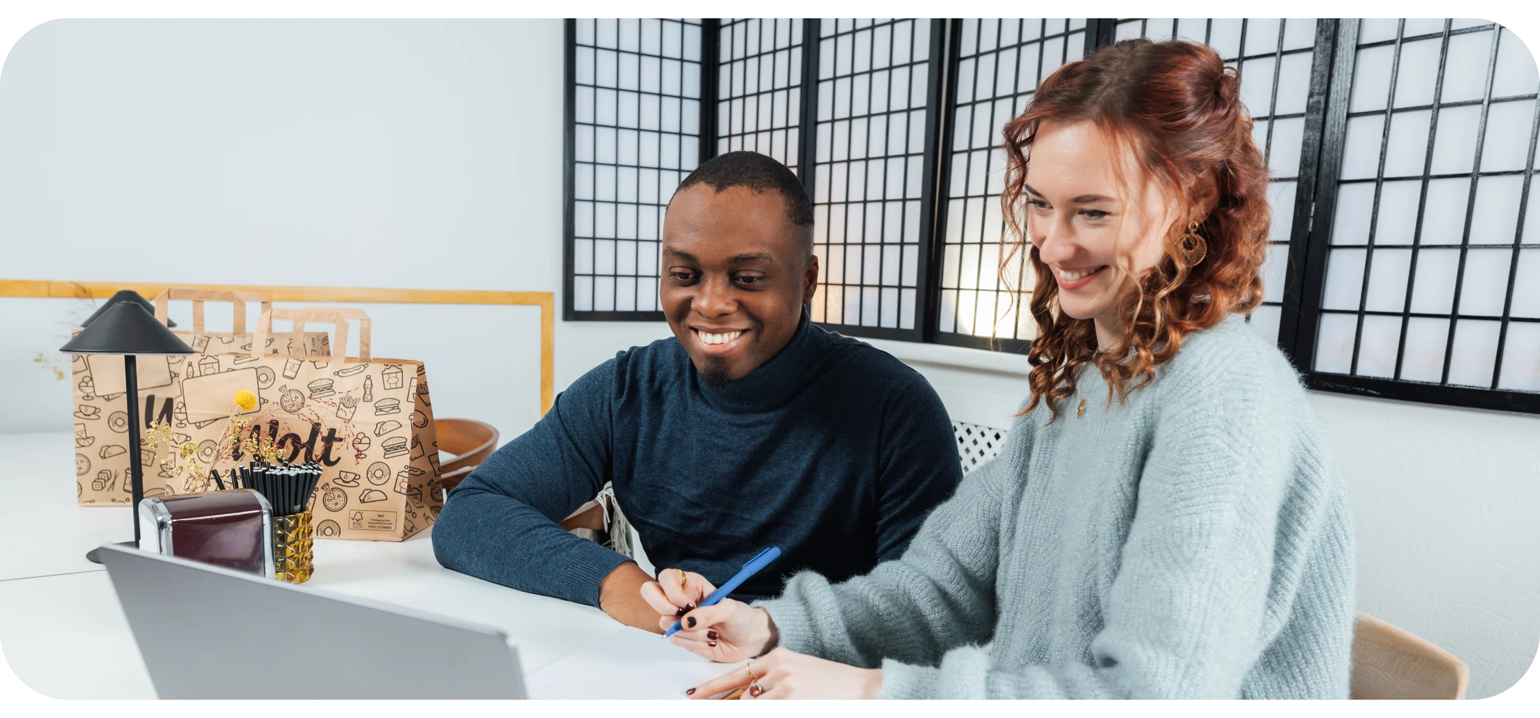 A man and woman sit at a desk smiling and writing on paper, with a laptop and paper Wolt bags nearby.