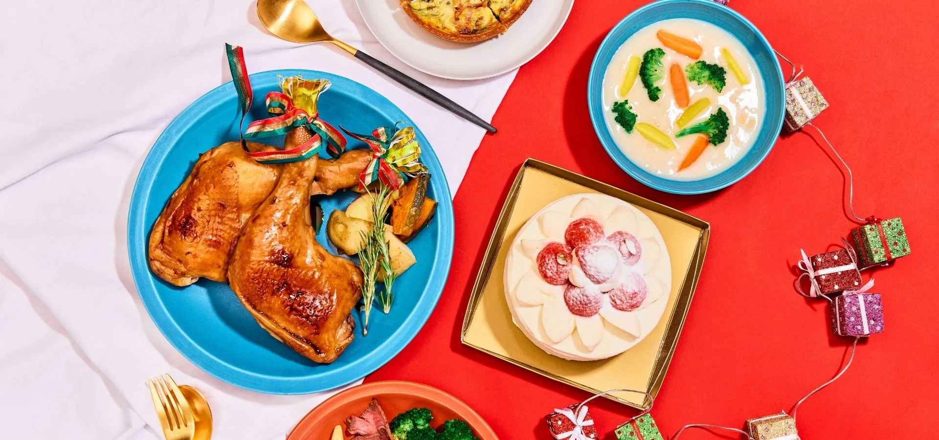 Hähnchenschenkel mit Schleife auf einem blauen Teller, eine cremige Gemüsesuppe, eine Erdbeer-Sahne-Torte und kleine verpackte Geschenke vor rotem Hintergrund