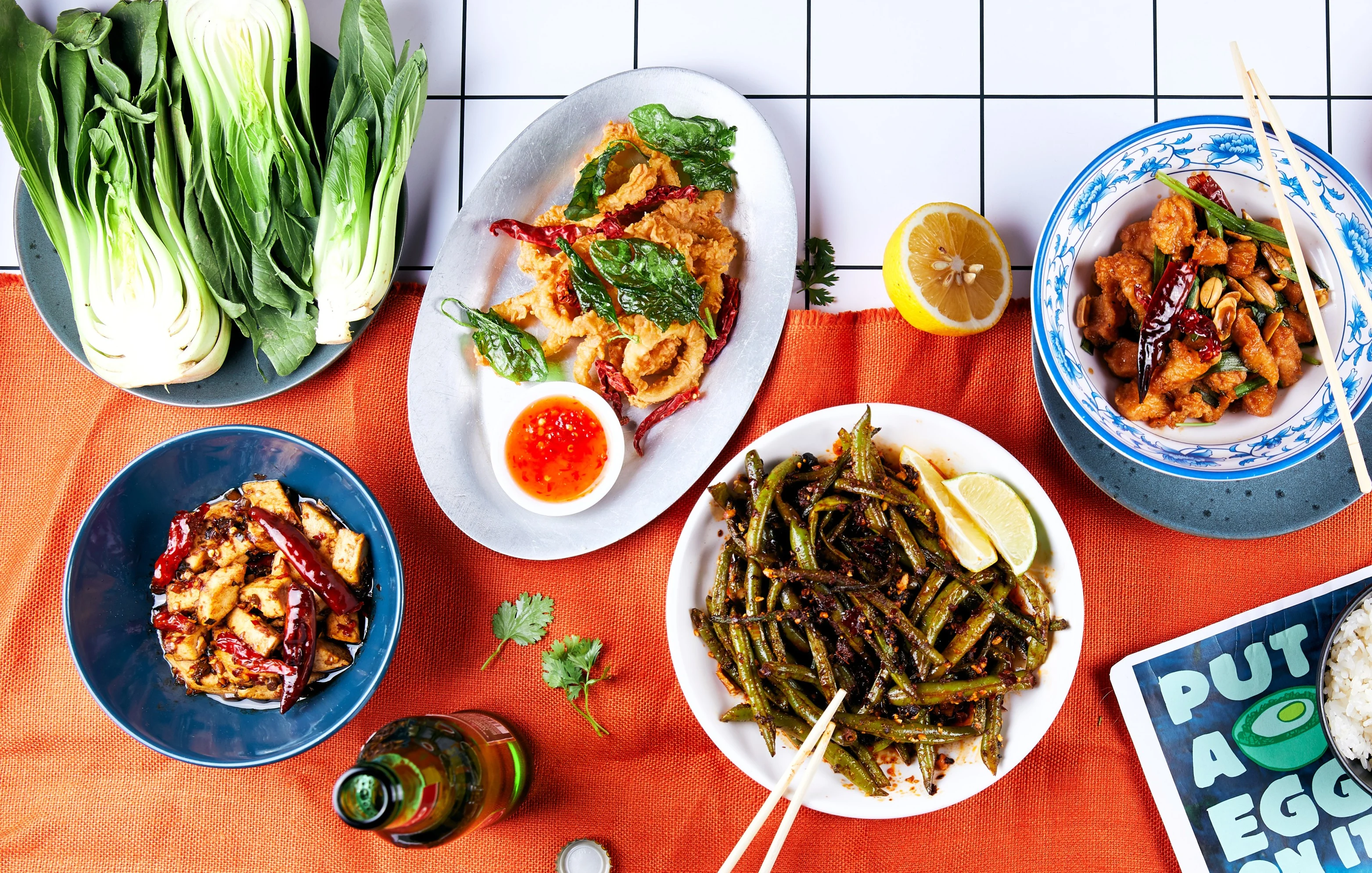 Verschiedene Gerichte auf einem orangefarbenen Tuch, mit gebratenen grünen Bohnen mit Limette, frittierten Tintenfischringen mit Chili, einem Hähnchengericht mit Erdnüssen und getrockneten Chilis, geschmortem Tofu mit Chili, Pak Choi und einer Flasche Bier