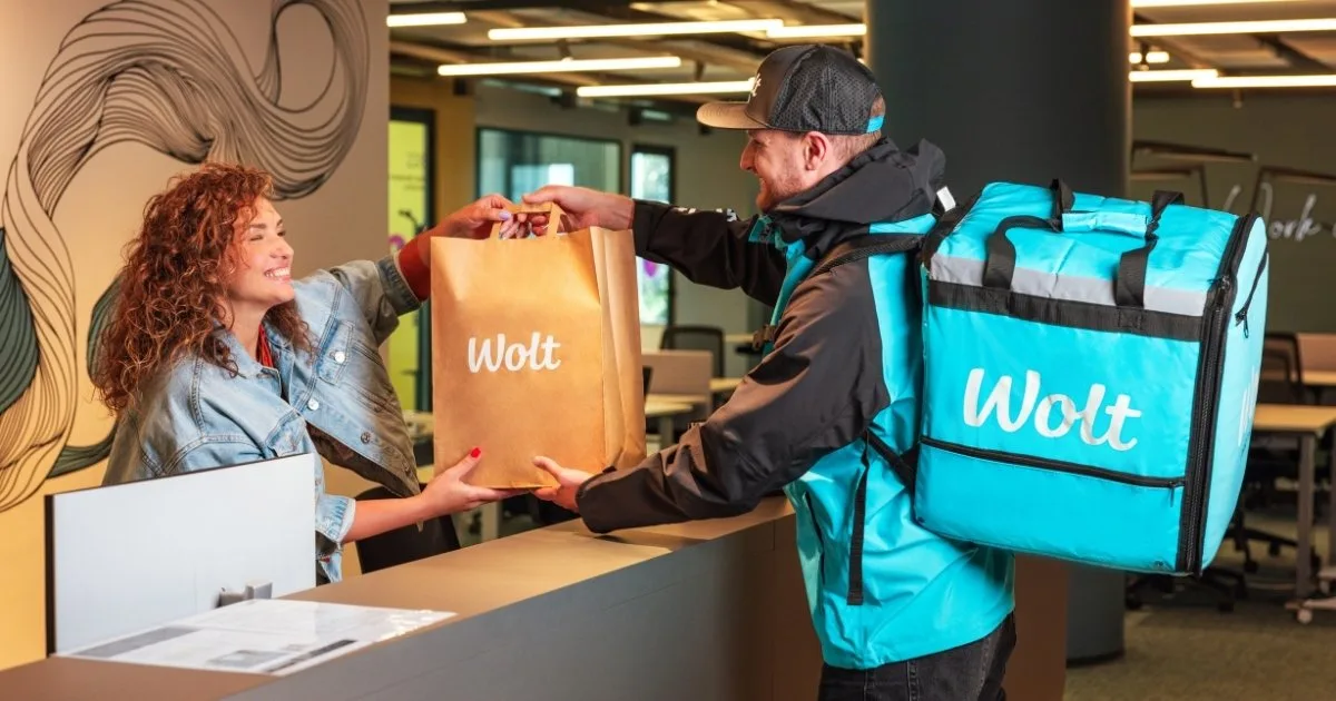 Ein Wolt-Kurier übergibt eine Liefertüte an eine lächelnde Kundin am Empfang – Liefer-App-Werbung führt direkt zu abgeschlossenen Bestellungen, anders als traditionelles Marketing