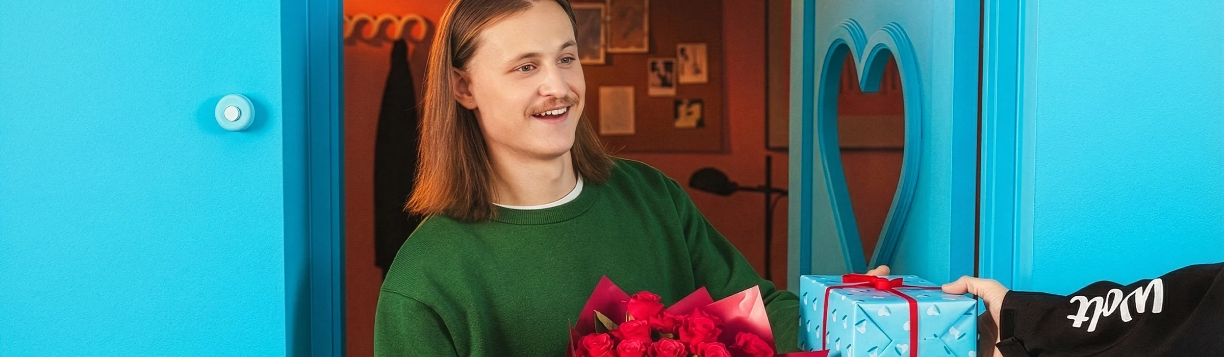 Person standing in a blue doorway, holding a bouquet of red roses while receiving a wrapped gift from a Wolt delivery courier.