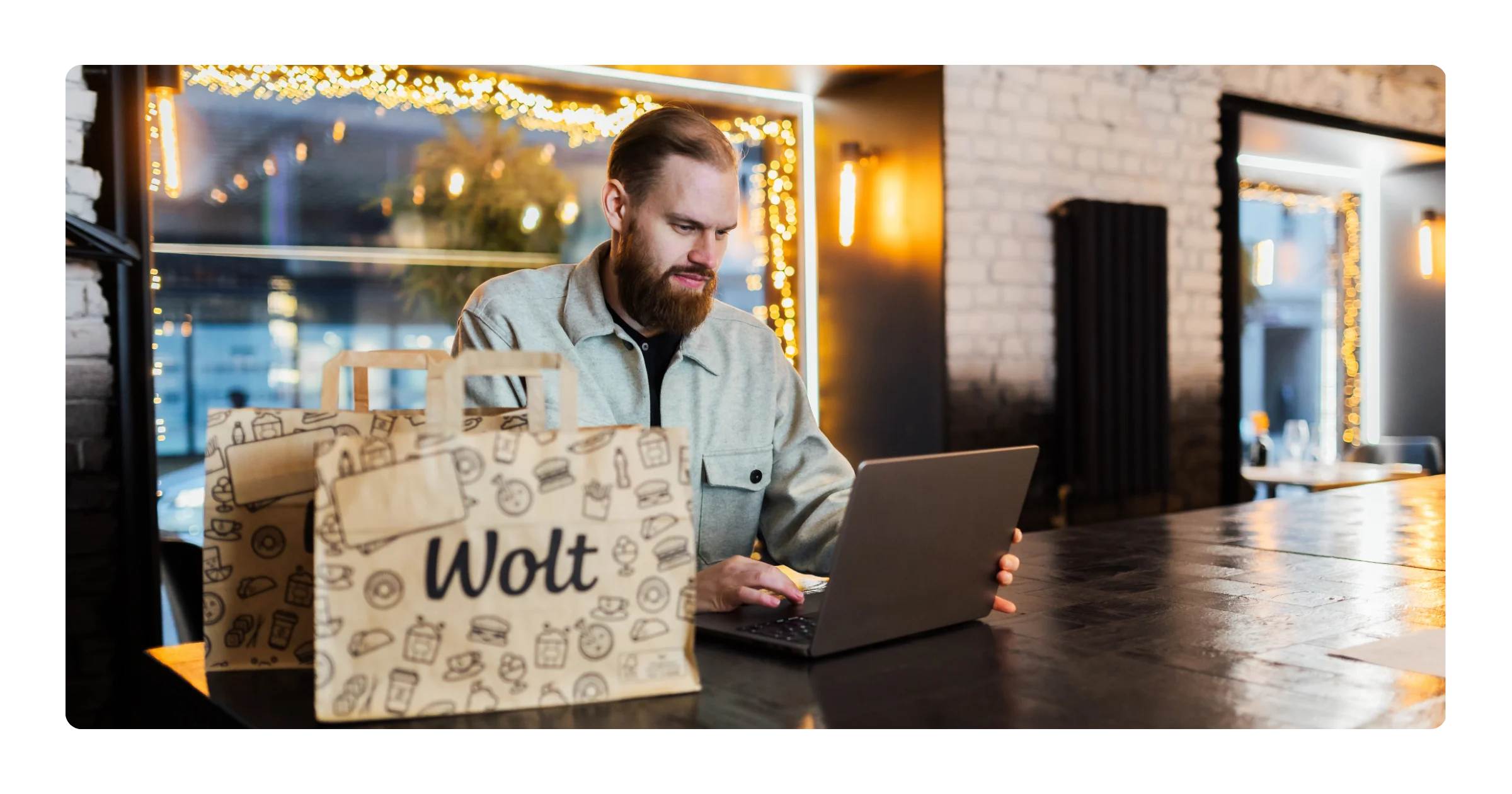 A bearded man works on a laptop at a restaurant table, with two paper Wolt delivery bags placed in front of him.
