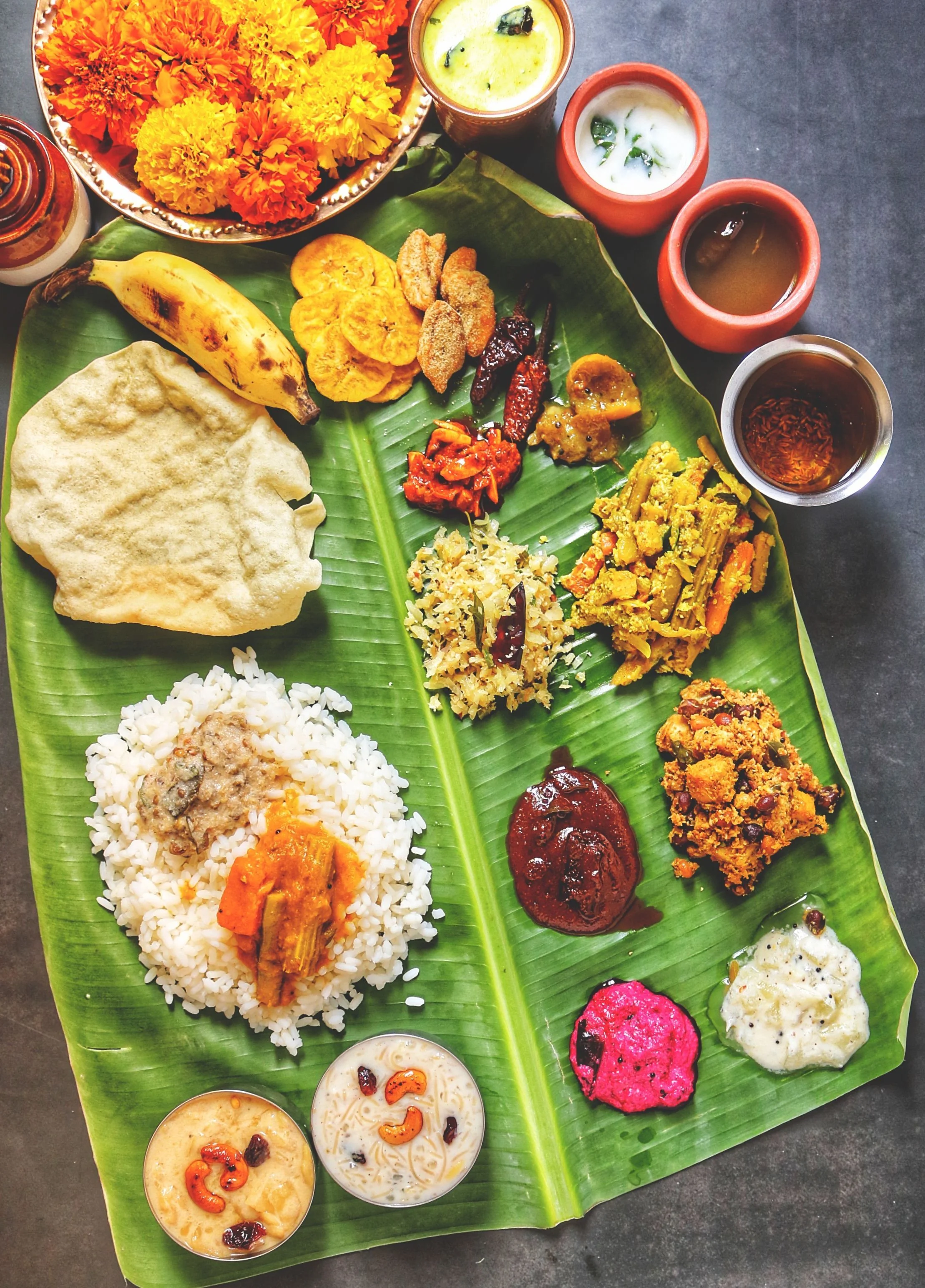 A South Indian sadya served on a banana leaf, with rice, papadum, plantain, banana chips, several vegetable curries and chutneys, pickles, and payasam desserts, alongside marigolds and pots of buttermilk and rasam