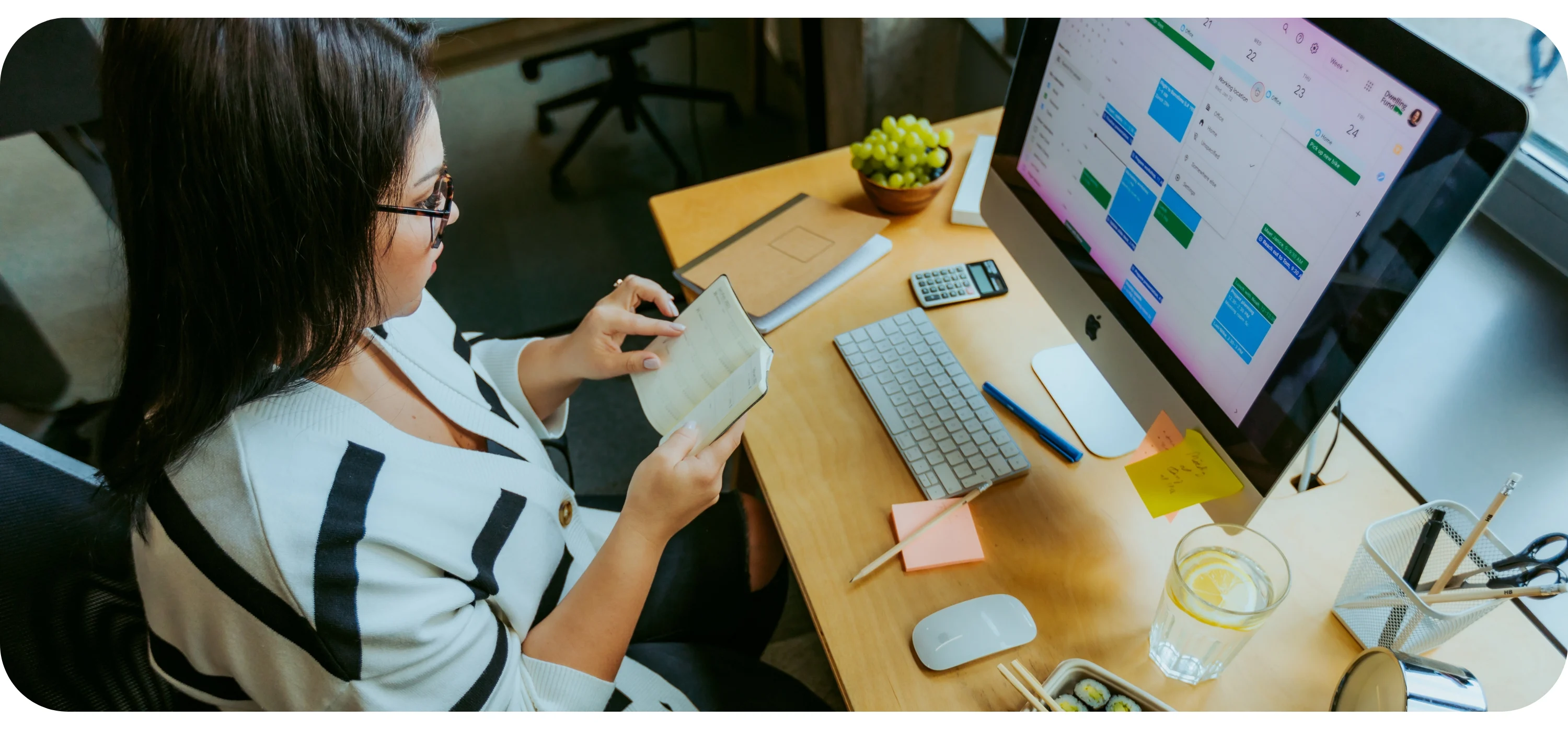 A woman in glasses reads a notebook at a desk with a computer showing a calendar