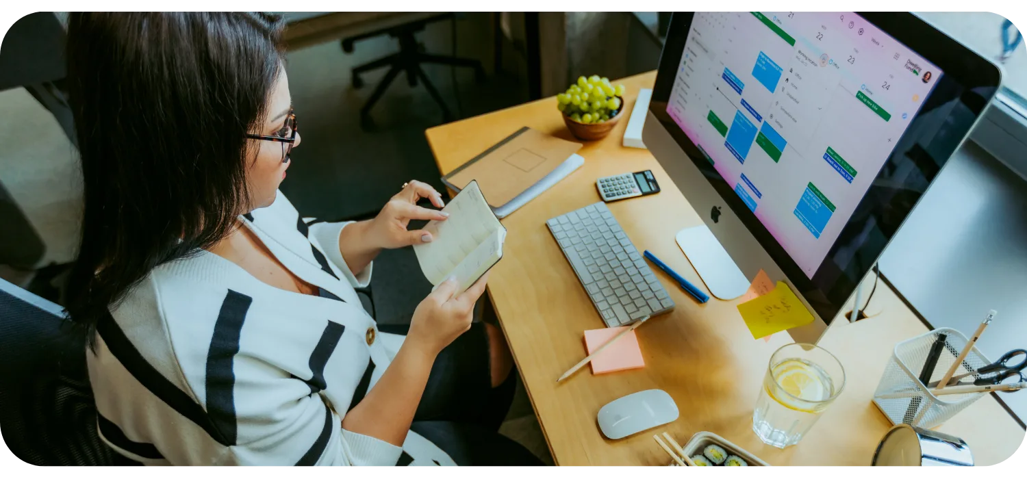 A woman in glasses reads a notebook at a desk with a computer showing a calendar