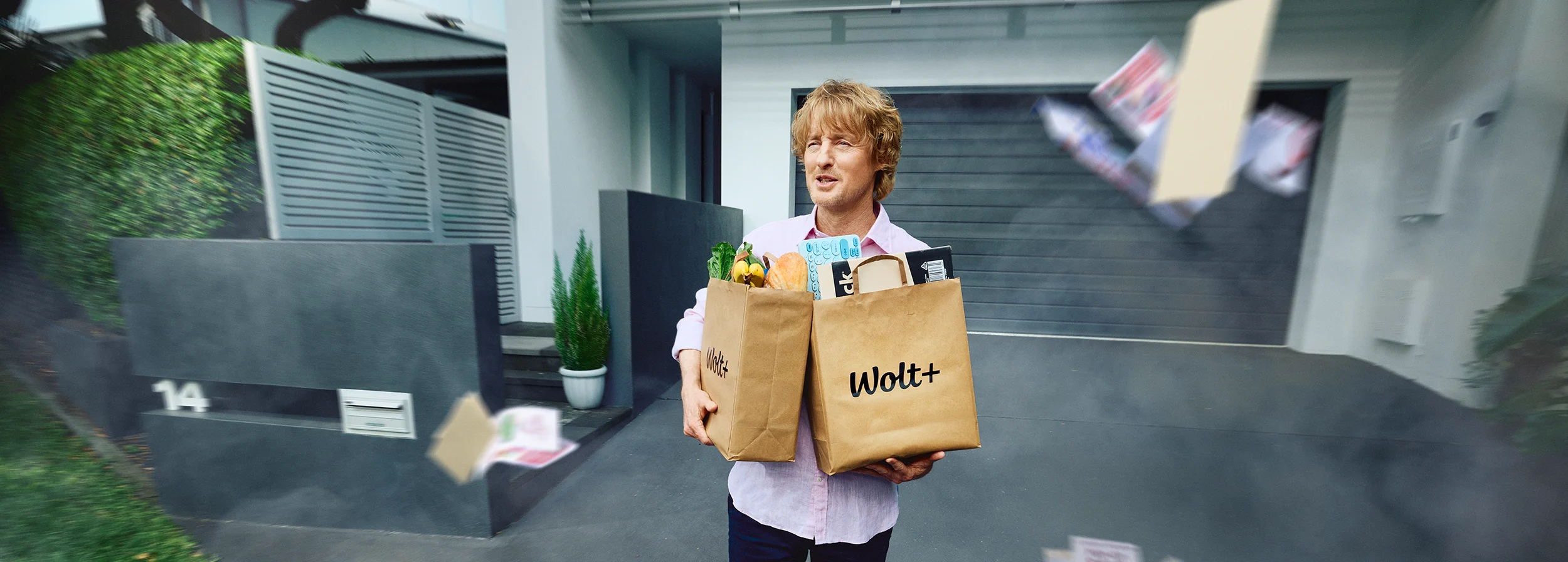 A person stood holding two Wolt+ paper bags with groceries and electronic products visible inside.