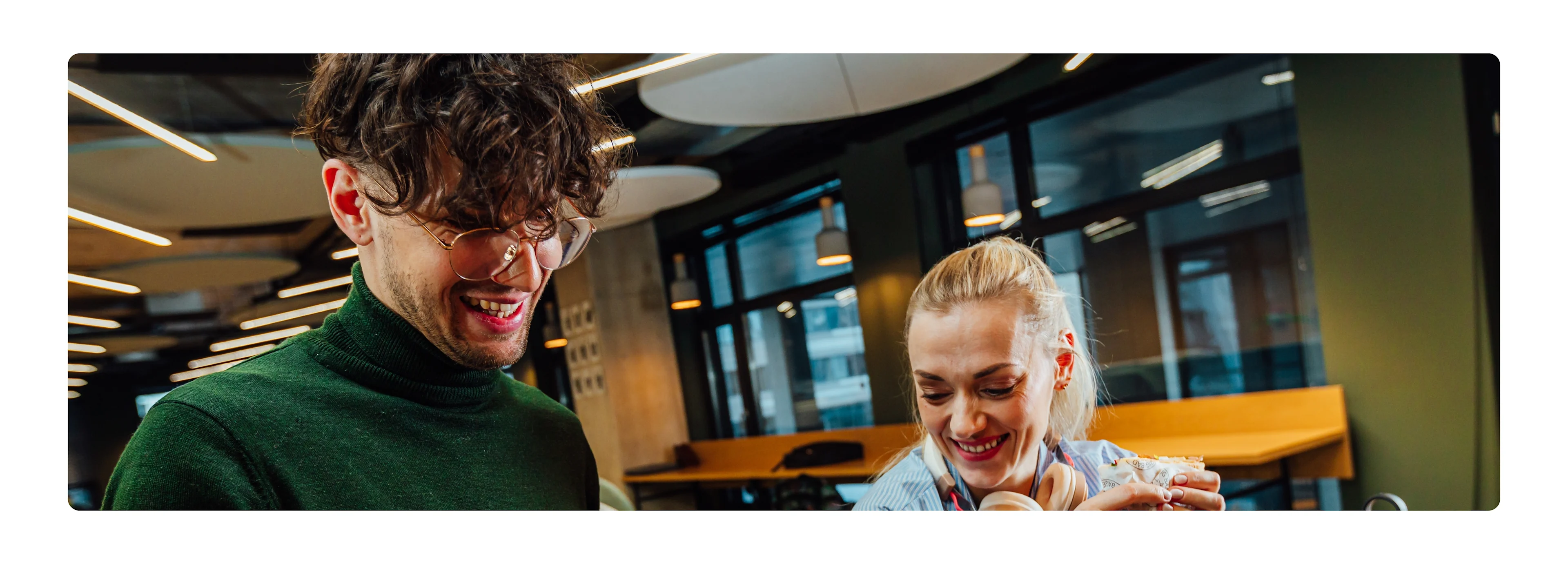 A smiling restaurant worker in glasses and a colleague holding a wrapped sandwich share a relaxed moment in a busy venue