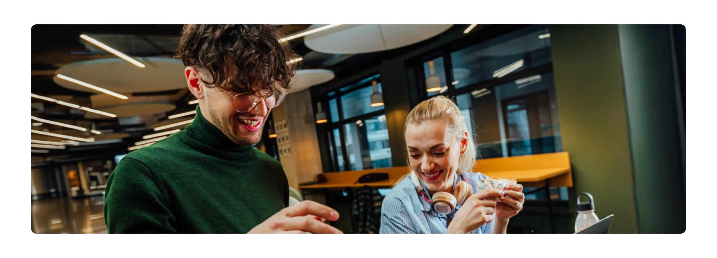 A smiling restaurant worker in glasses and a colleague holding a wrapped sandwich share a relaxed moment in a busy venue