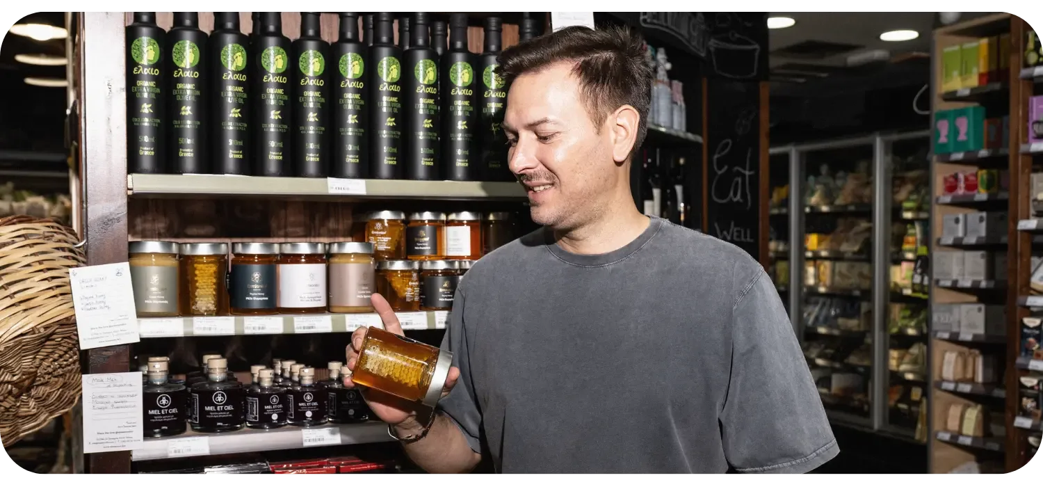 A man inside the 4 Seasons Bio grocery store in Athens smiles while holding a jar of honey with honeycomb, standing in front of shelves stocked with organic olive oil and other local products.