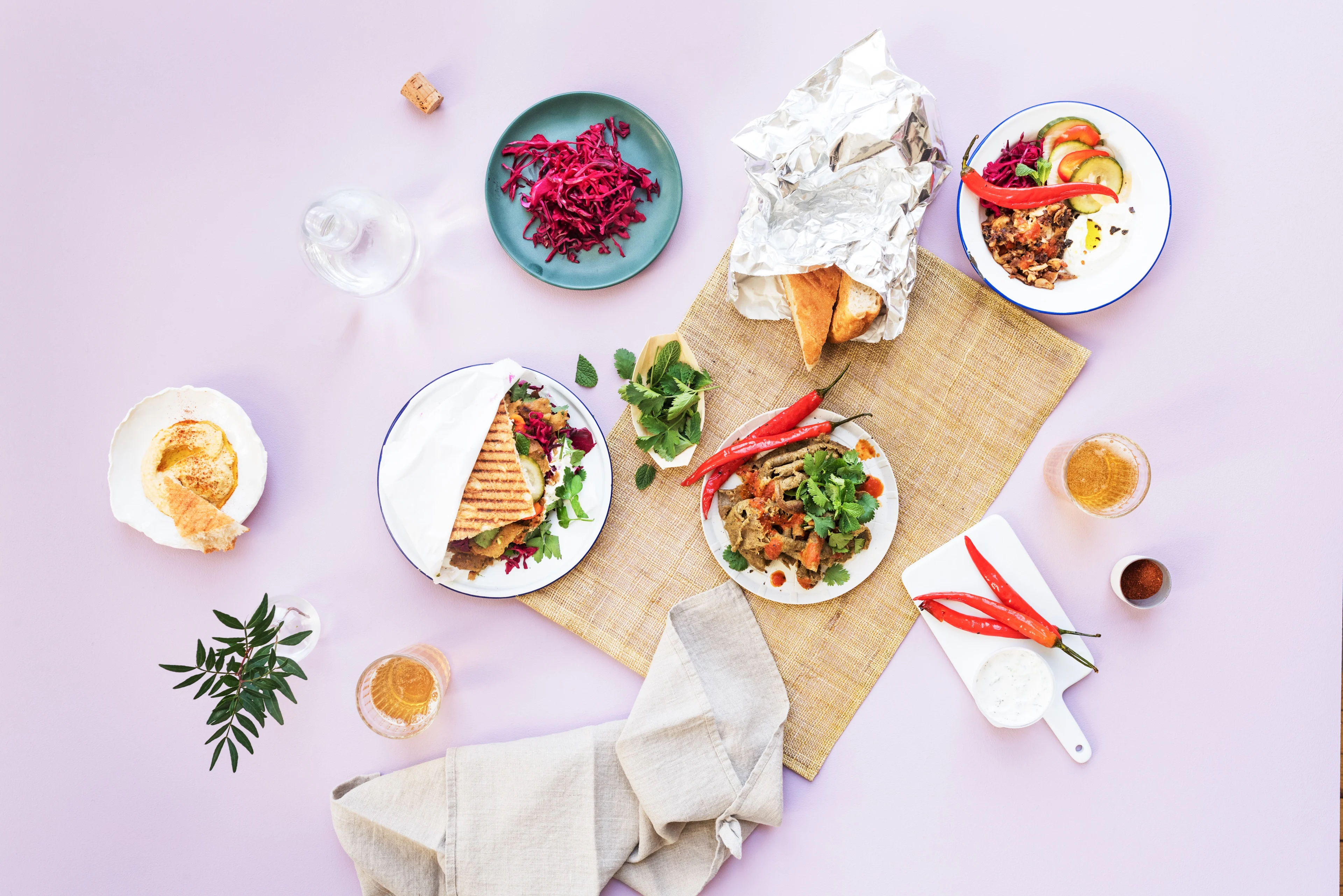 A Turkish spread on a pink background, with döner kebab plates, a grilled wrap, red cabbage salad, hummus, bread, fresh chillies, herbs and glasses of beer