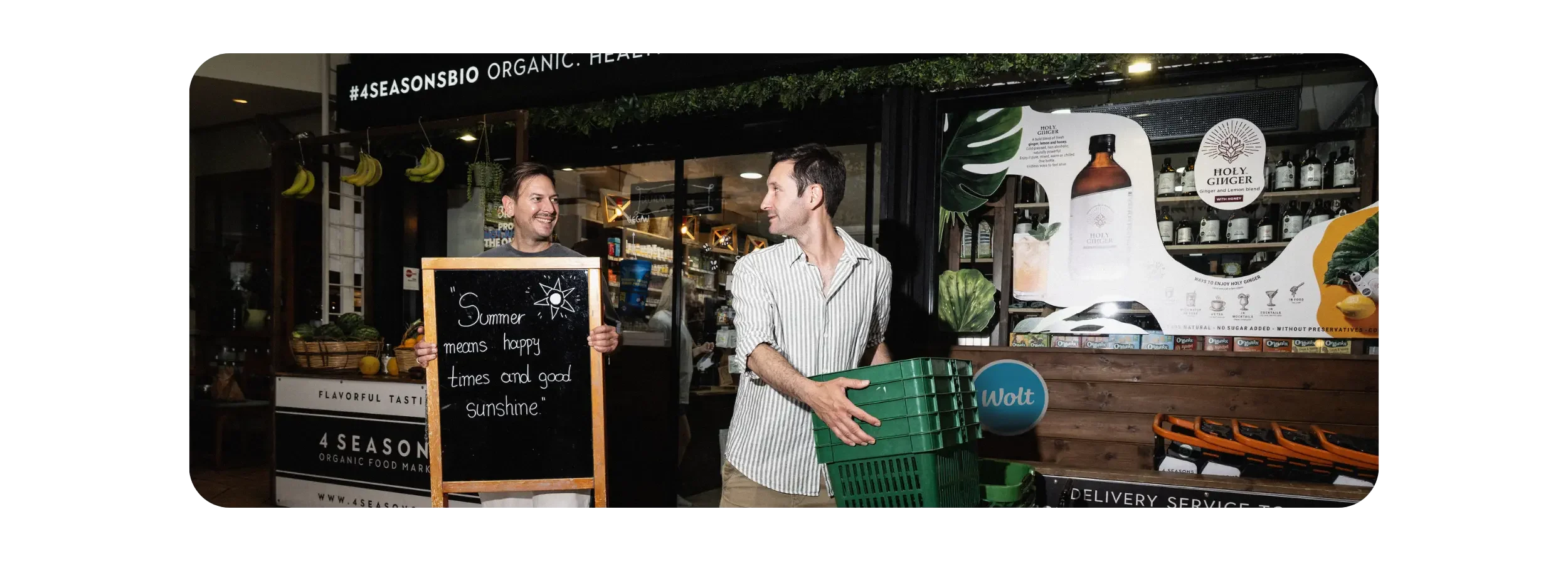 Two brothers outside their 4 Seasons Bio grocery store in Athens, smiling as one carries a sign reading “Summer means happy times and good sunshine,” while the other holds a stack of green shopping baskets.