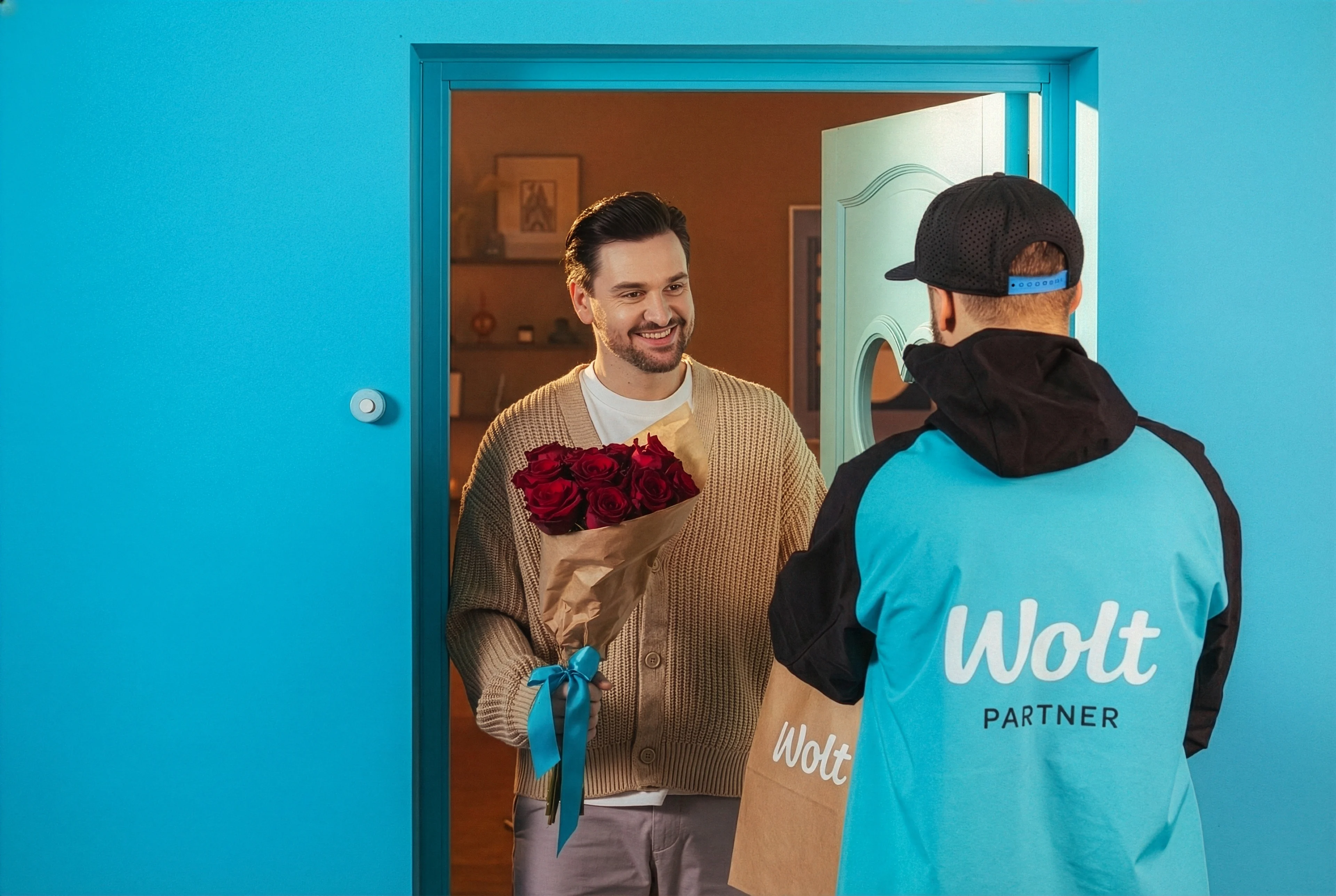 Courier wearing a Wolt jacket hands a bouquet of red roses to a smiling man at his front door.