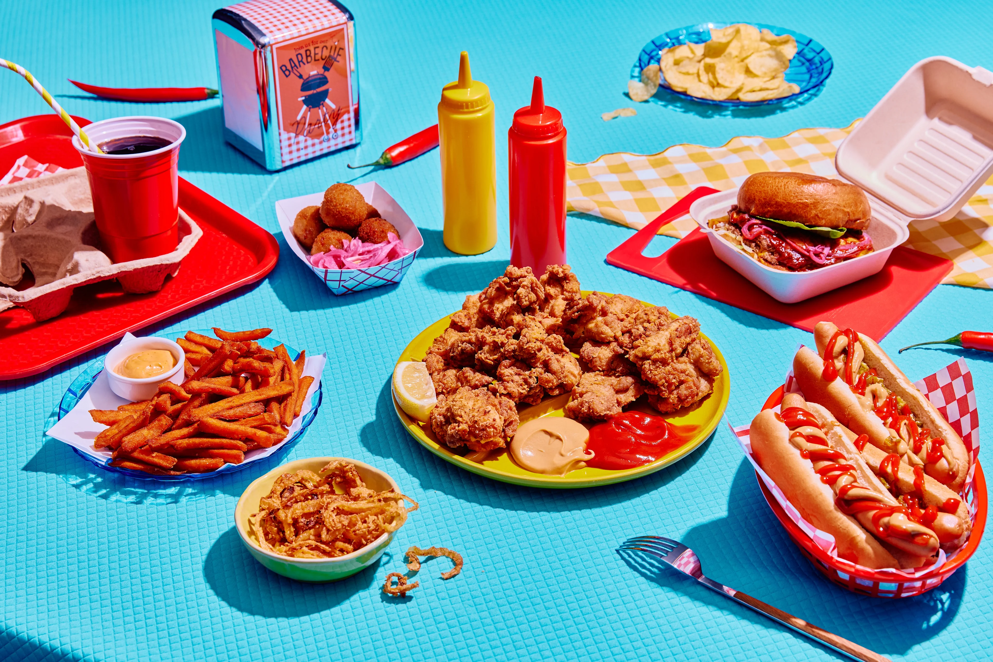 Ein amerikanisches Diner-Arrangement vor blauem Hintergrund, mit frittiertem Hähnchen, einem Hot Dog mit Ketchup und Senf, einem Burger in einer Takeaway-Box, Süßkartoffel-Pommes, frittierten Zwiebeln, Kartoffelchips, einer Cola im roten Becher sowie Ketchup- und Senfflaschen