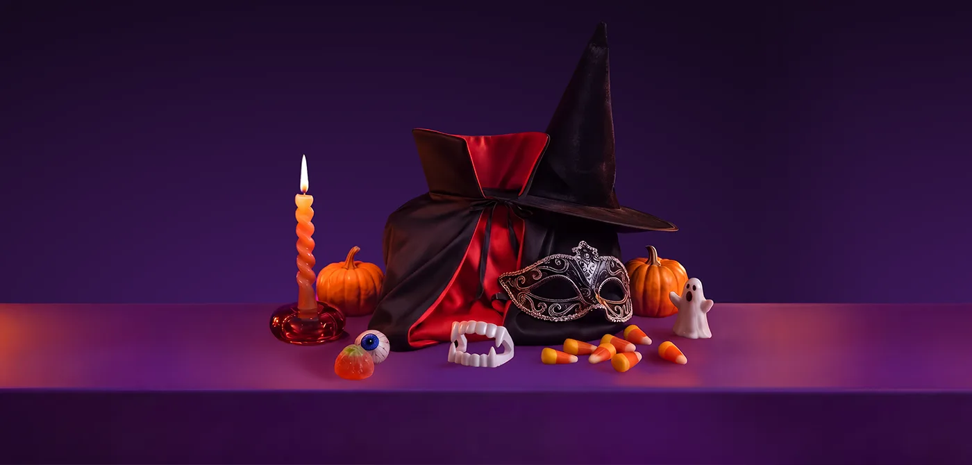 A black and red vampire cape with a witch hat, a silver masquerade mask, plastic vampire fangs, a glow-in-the-dark eyeball, candy corn sweets, a small pumpkin and a ghost figurine, beside a lit spiral candle, on a purple surface, representing Halloween costume essentials delivered with Wolt.
