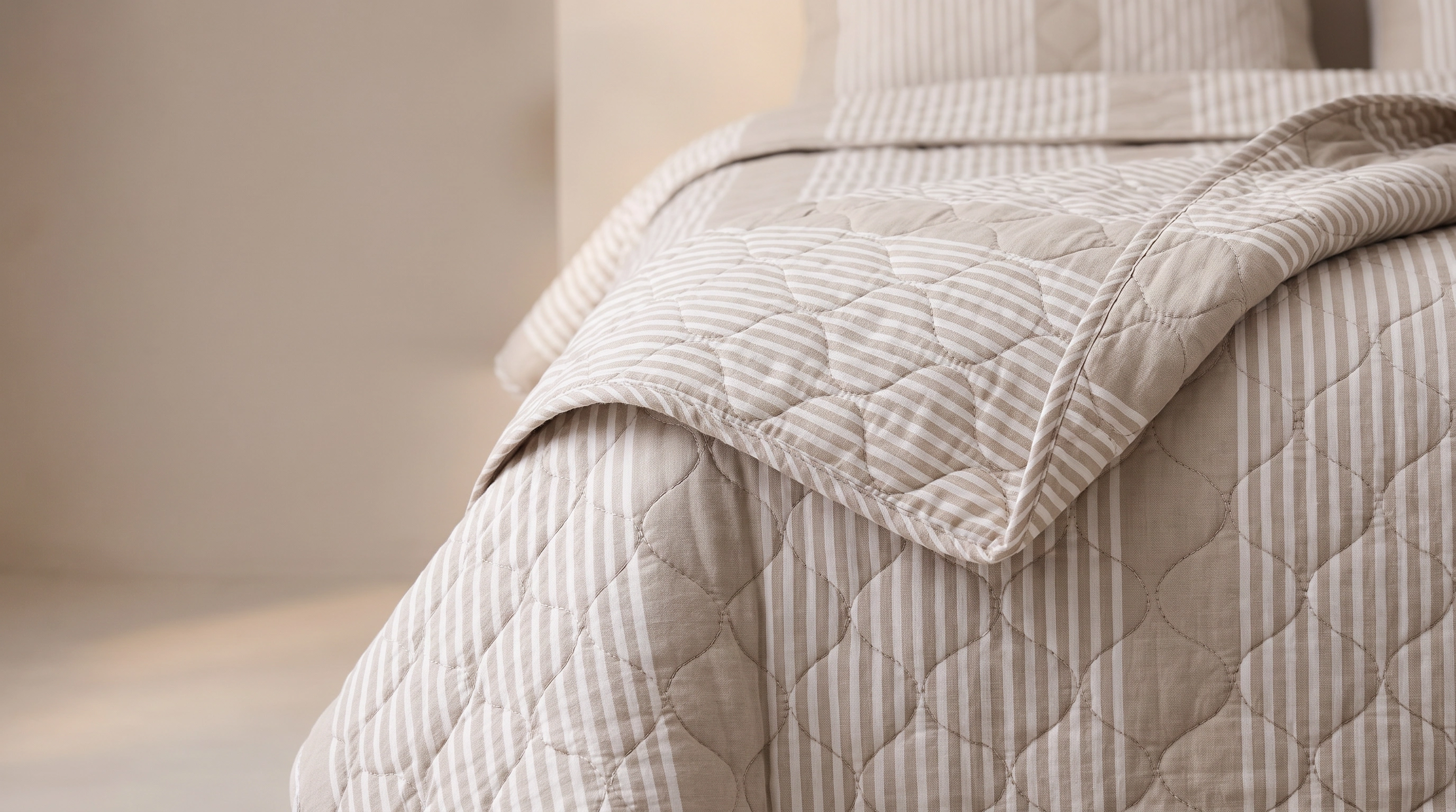 A beige striped quilt folded back at the corner of a made bed, its scalloped quilting catching the light