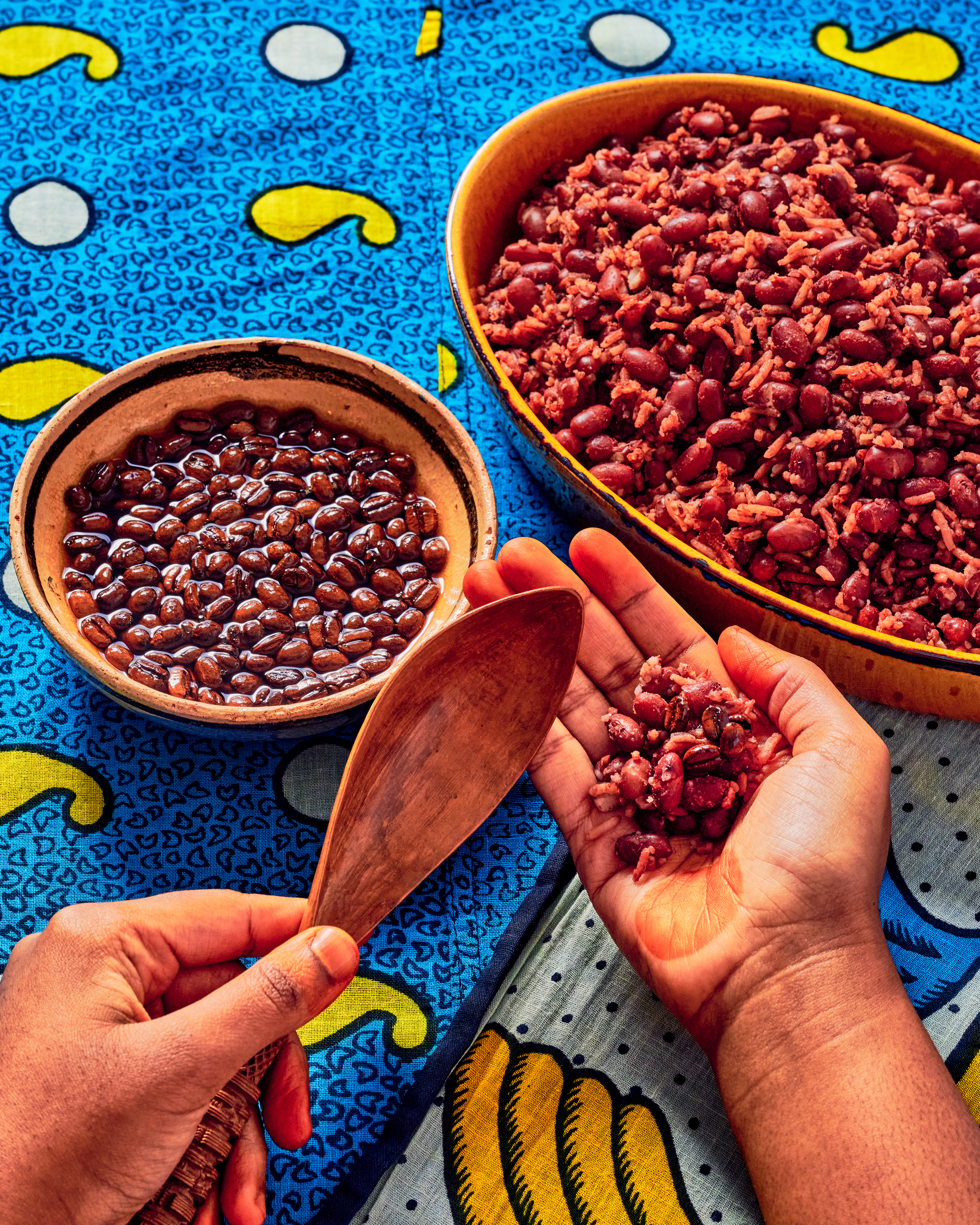 Hands serving red beans and rice from colorful bowls on a vibrant blue patterned tablecloth, with a wooden spoon.