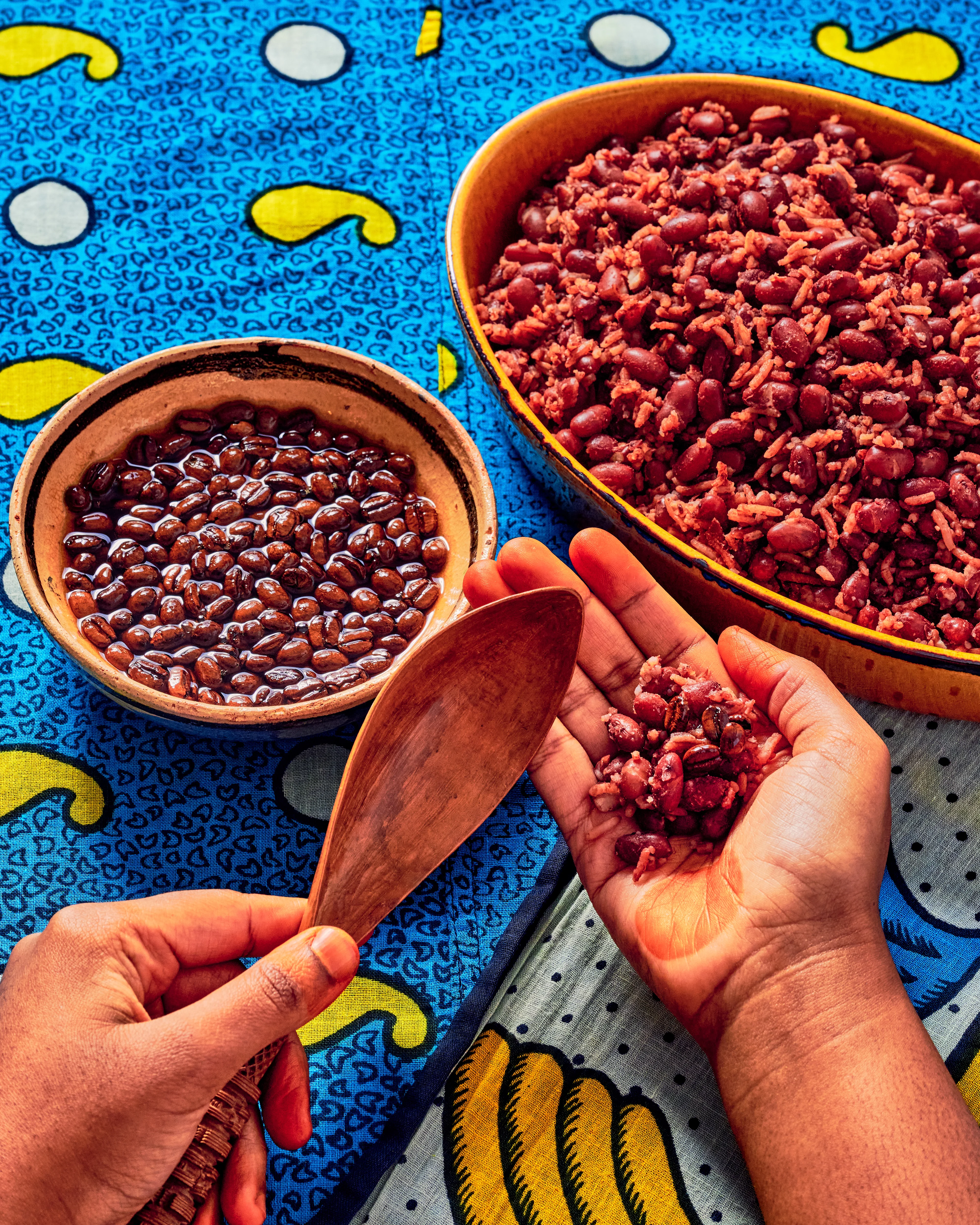 Hands serving red beans and rice from colorful bowls on a vibrant blue patterned tablecloth, with a wooden spoon.