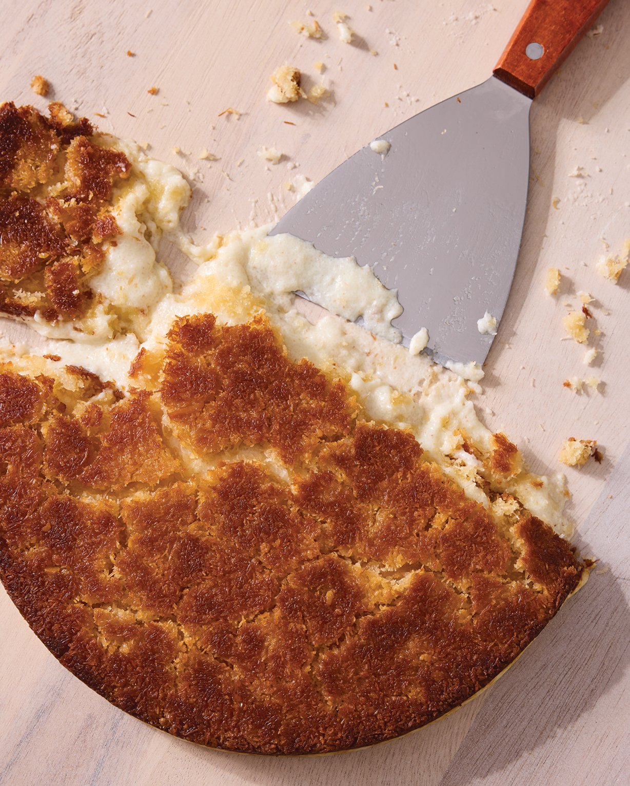 Golden-brown baked dessert with creamy filling being served with a metal spatula on a light wooden surface.